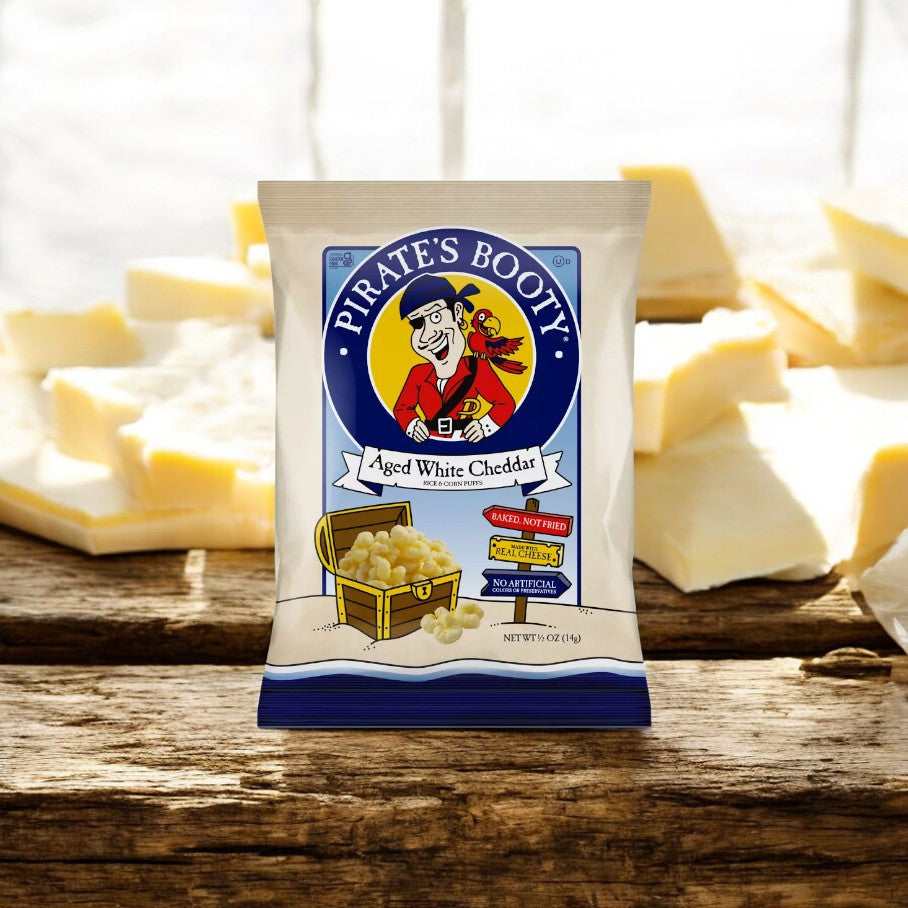 A bag of Pirate's Booty Aged White Cheddar Puffs .5 oz is placed on a wooden surface, surrounded by blocks of cheese in the background.