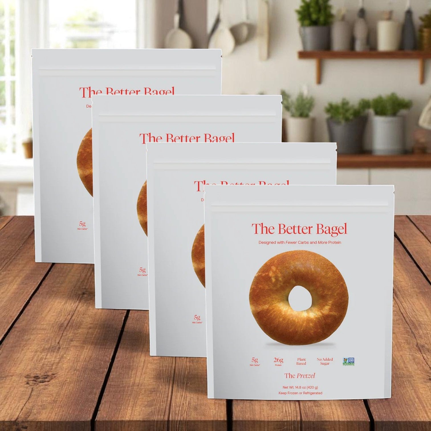 Four bags of BetterBrand’s The Better Bagel – Pretzel Style (16 bagels total) are shown on a wooden kitchen surface, with plants and kitchenware in the background. Now in stock and ready to ship!.