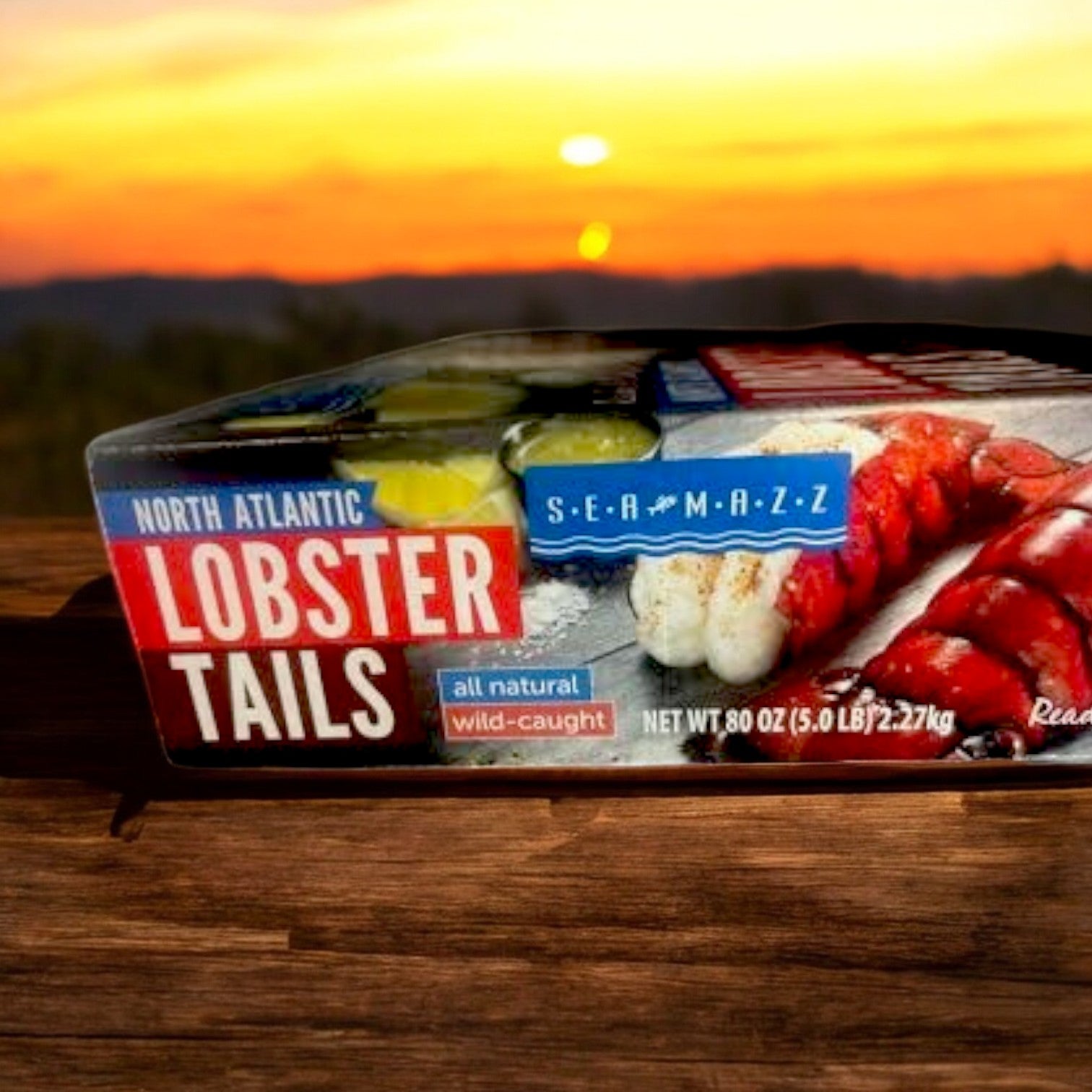 A 5-pound box of Seamazz North Atlantic Lobster Tails, 4 oz each, labeled "all natural" and ready to cook, rests on a wooden surface with a blurred outdoor sunset as the backdrop.