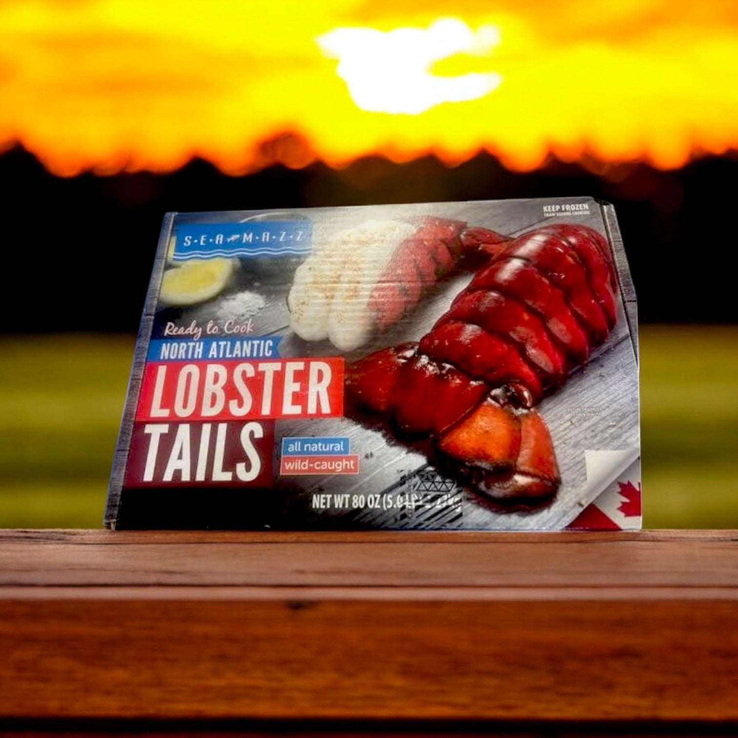 A box of Seamazz Ready to Cook North Atlantic Lobster Tails sourced sustainably, weighing 5 pounds, rests on a wooden surface, framed by a sunset.