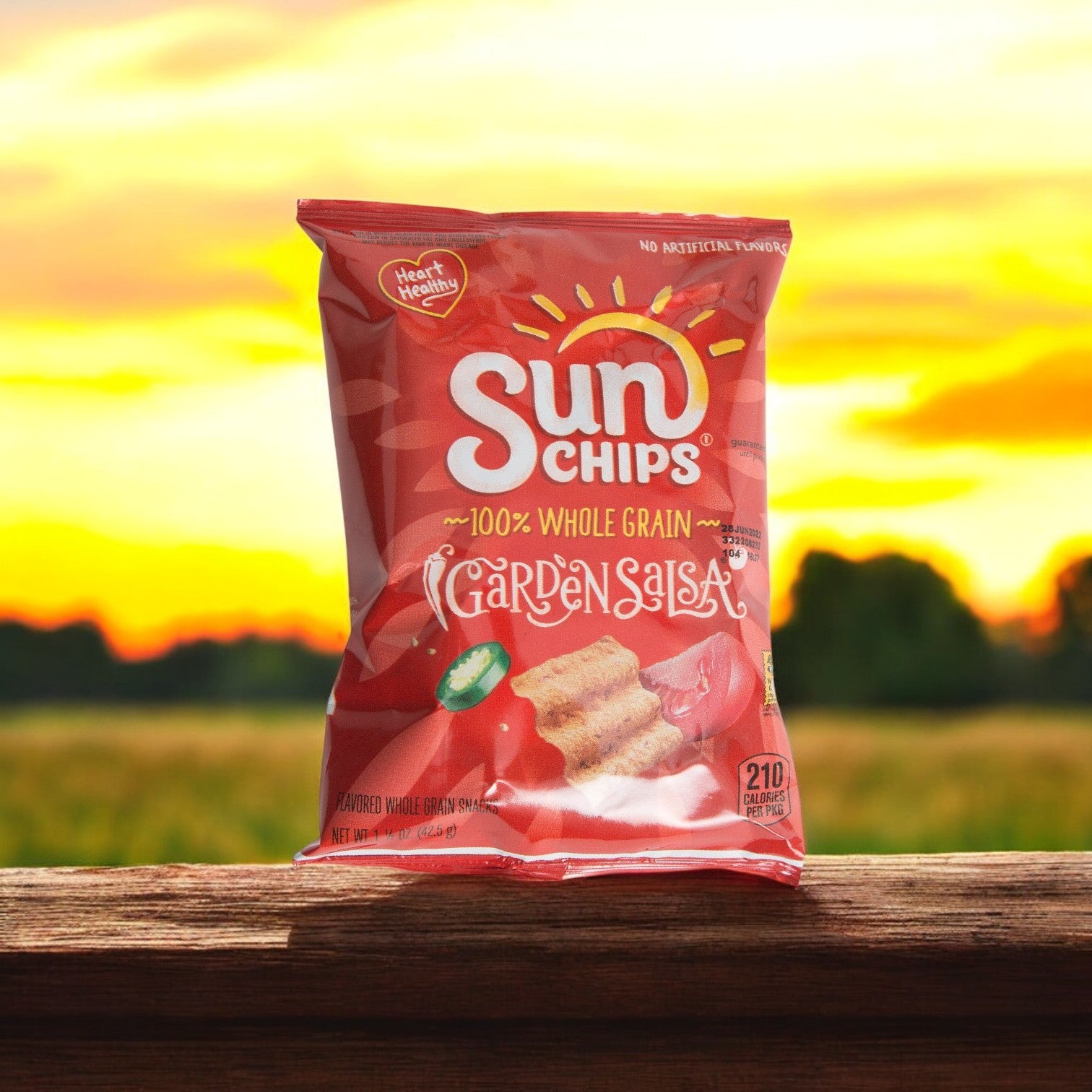 A 1.5 oz Sunchips Multigrain Garden Salsa bag from Sun Chips lies on a wooden surface, embodying whole grains with a lively sunset backdrop.