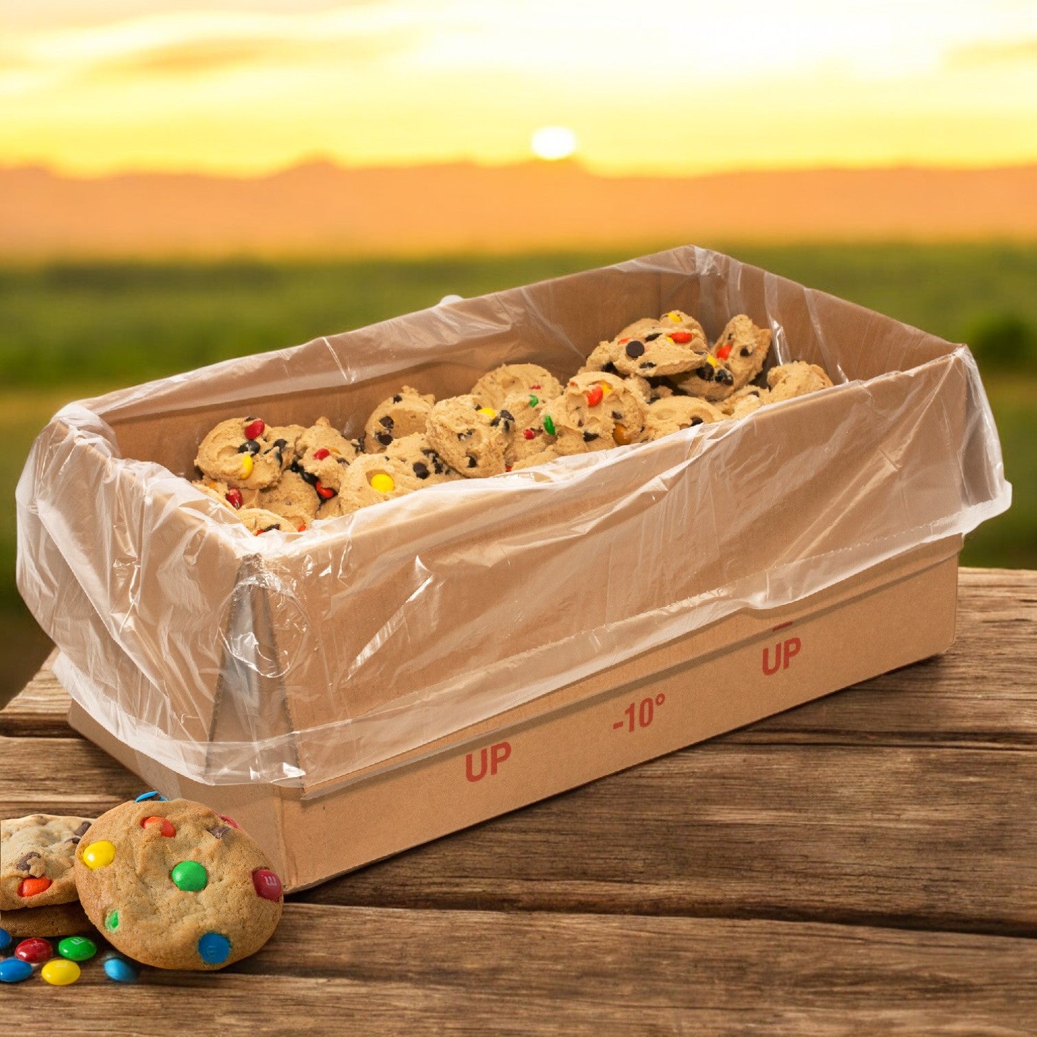 A cardboard box lined with plastic contains David's Cookies Dough Cookie - Chocolate Chip M&M topped with colorful candies, resting on a wooden table at sunset.