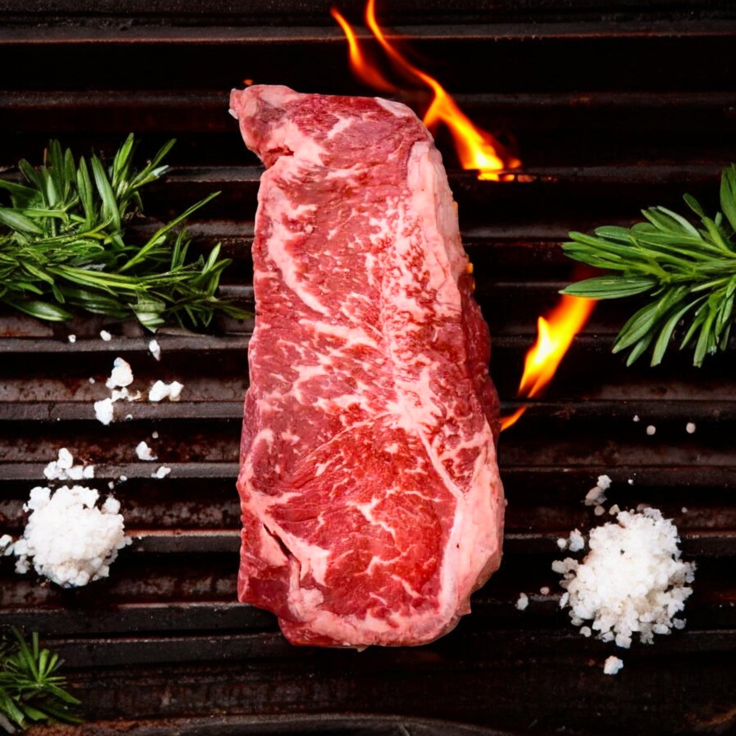 The 10 oz Mino Prime Steaks New York Strip Steak sizzles on the grill as flames dance around its marbled surface, enhanced by rosemary sprigs and coarse salt.