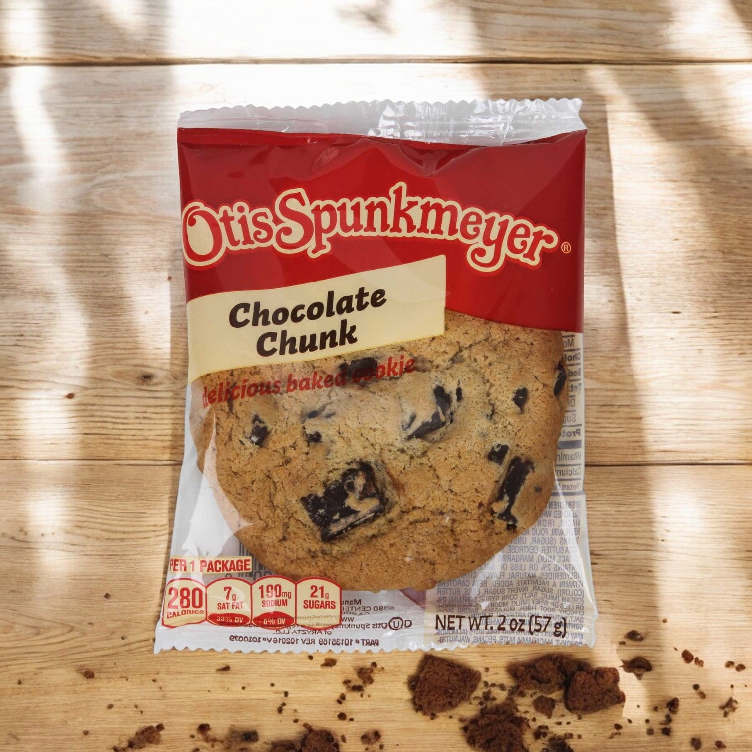 An Otis Spunkmeyer Individually Wrapped Chocolate Chip Cookie, 2 oz., rests enticingly on a wooden surface with chocolate chips peeking through. Cookie crumbs scatter around, hinting at its delicious texture.