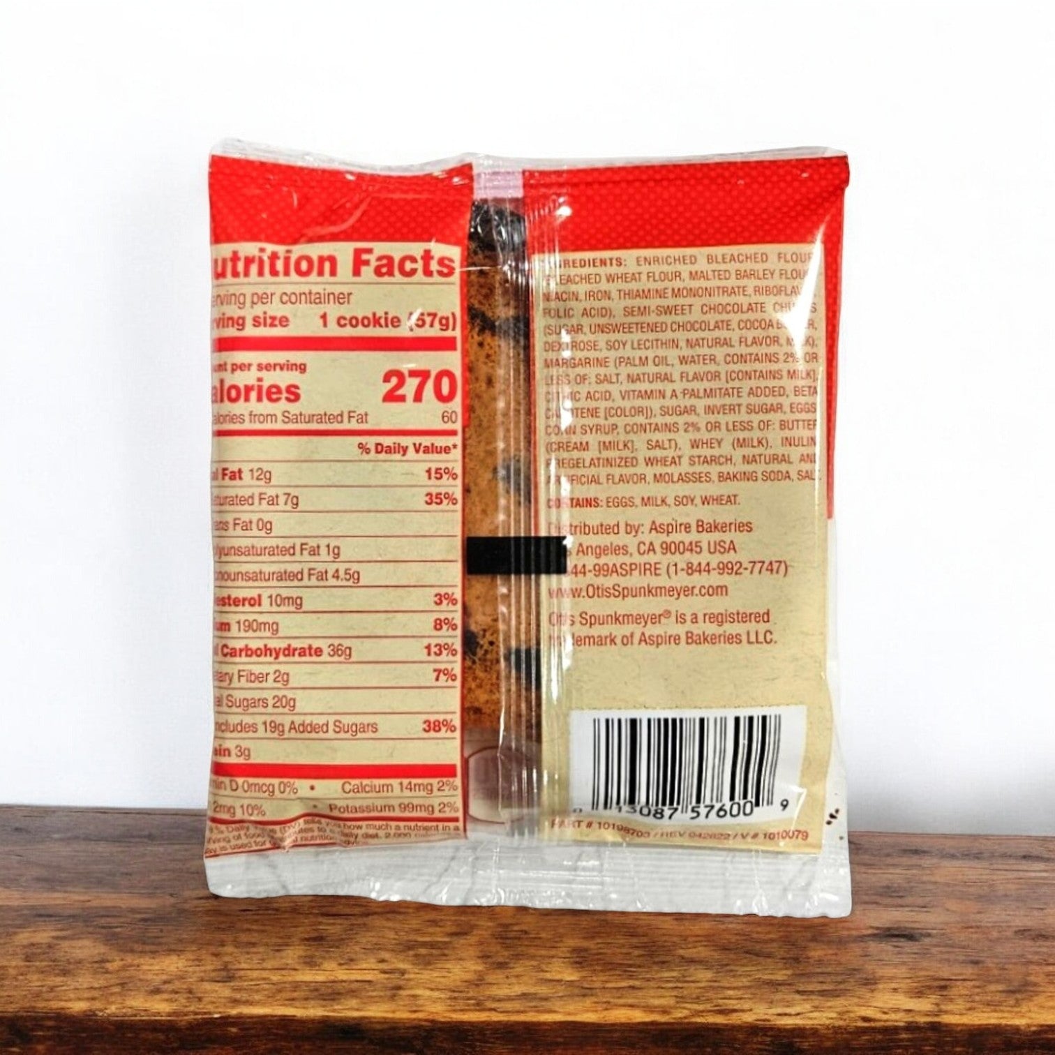 Rear view of the Otis Spunkmeyer Individually Wrapped Chocolate Chip Cookies, 2 oz. - 1 Count package displaying nutrition facts and ingredients list. The partially transparent wrapper reveals the cookie with chocolate chips inside. The individually wrapped cookie is placed on a wooden surface.