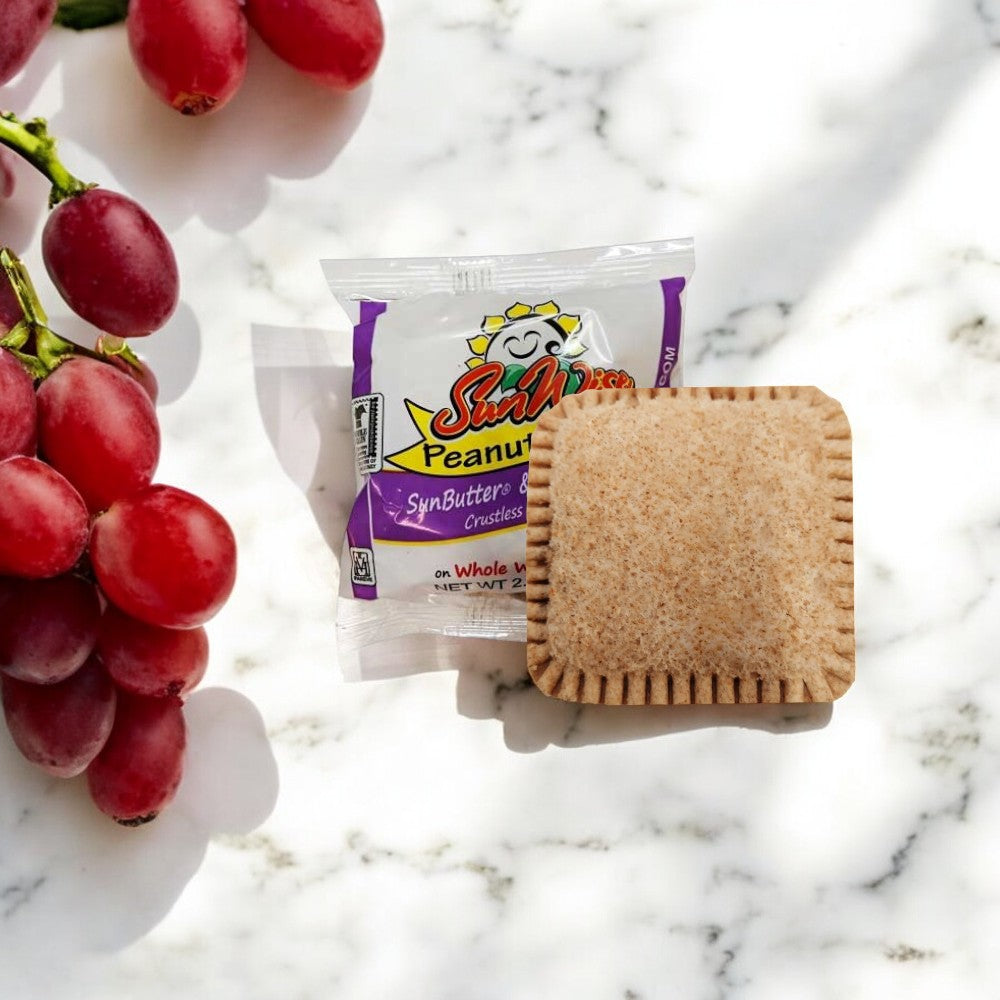 A package of Sunwise Grape Jelly Sandwiches from a case containing 96 sandwiches sits next to an unpackaged, nut-free sandwich on a marble surface, accompanied by a bunch of red grapes.