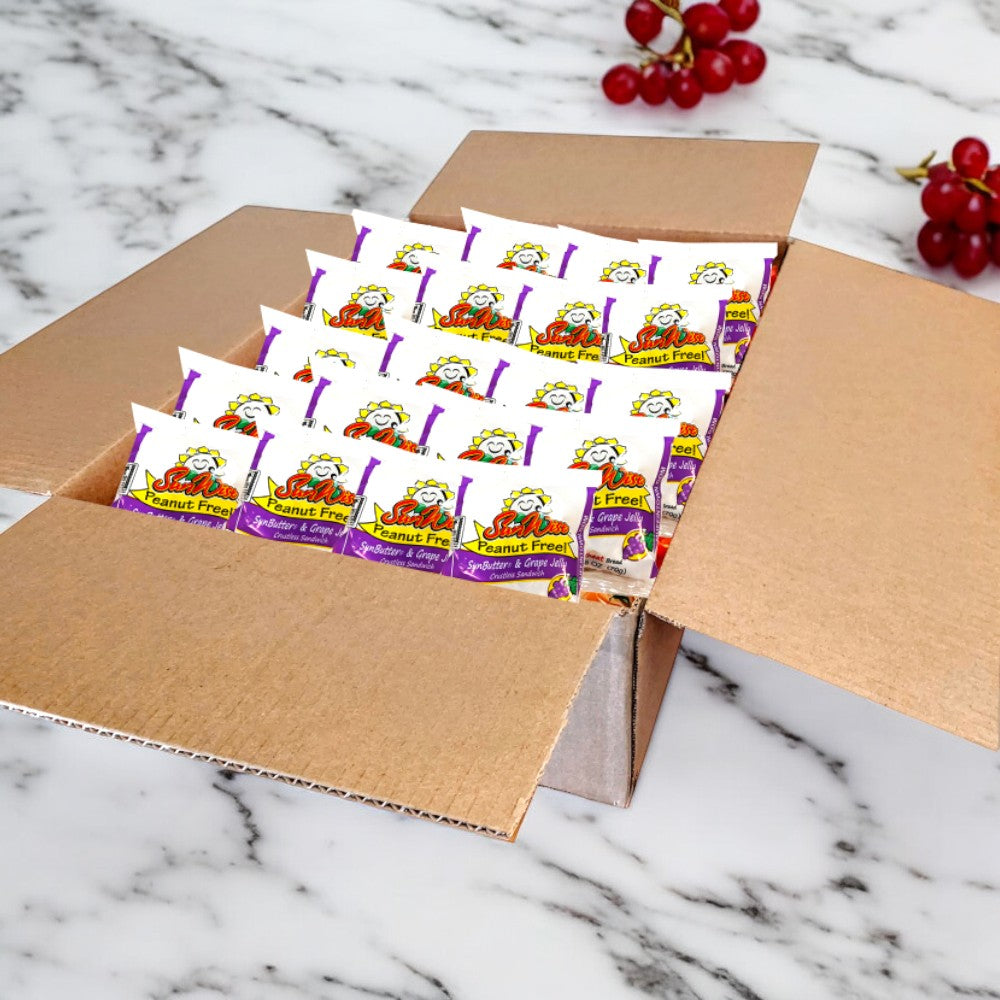 A cardboard box rests on a marble surface, filled with multiple packs of individually wrapped and "nut-free" SunWise Grape Jelly Sandwiches from a case of 96 by Sunwise. In the background, a small bunch of red grapes can be seen.