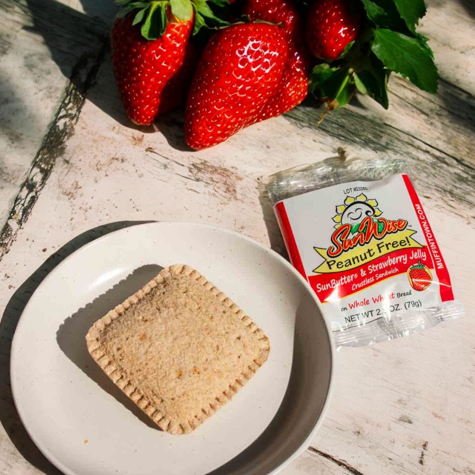 A wooden surface holds a plate featuring a sealed Sunwise Sunbutter and Strawberry Jelly Sandwich, beside fresh strawberries, and accompanied by a packet labeled Sunwise.