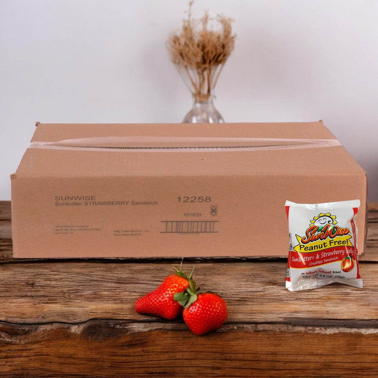 A cardboard box labeled "Sunwise - 96 Case-SunWise SunButter and Strawberry Jelly Sandwich," with a small packaged sandwich and several strawberries placed in front of it on a wooden surface.