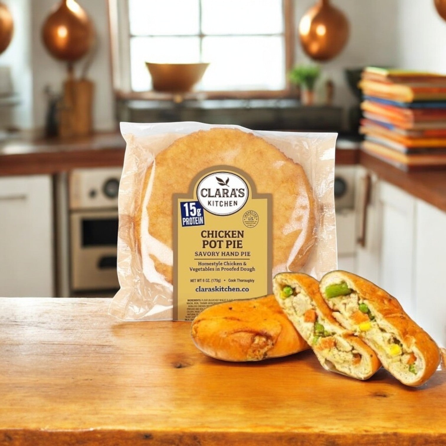 A Clara’s Kitchen Hand-Held Chicken Pot Pie, 6 oz., with its filling visible in a sliced portion, sits on a kitchen counter with a stove and bowls blurred in the background.