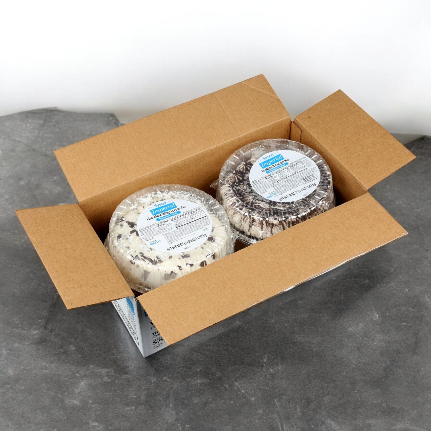 An open cardboard box shows two sealed circular cakes with Sysco labels, each a Sysco Imperial Assorted Cream Pie, promising creamy fillings and decadent desserts.