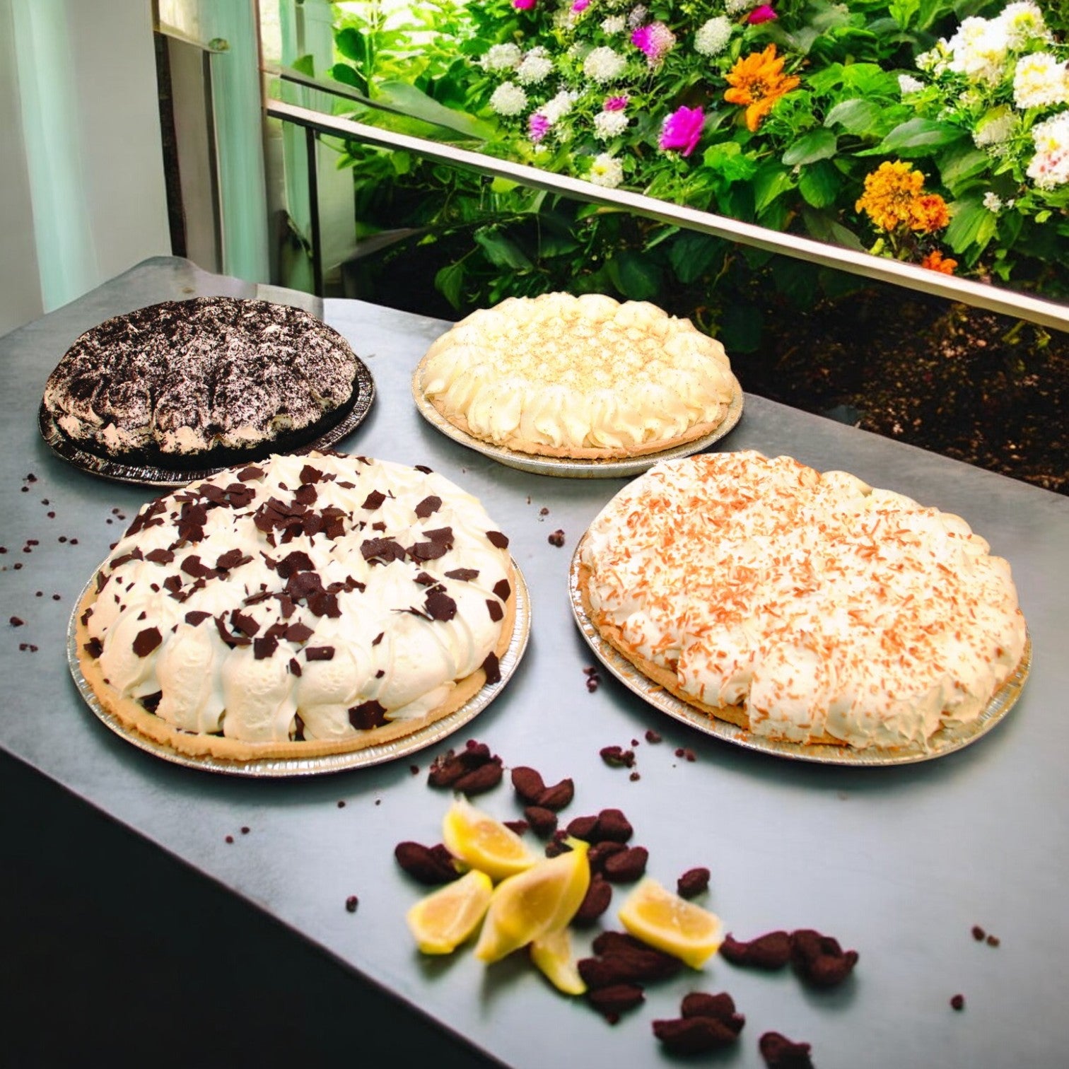 Four Assorted Cream Pies by Sysco Imperial, thaw and serve (4/10"), feature creamy fillings topped with chocolate shavings and nuts, elegantly placed on a metal surface with lemon wedges, chocolate pieces, and a backdrop of colorful flowers for added charm.