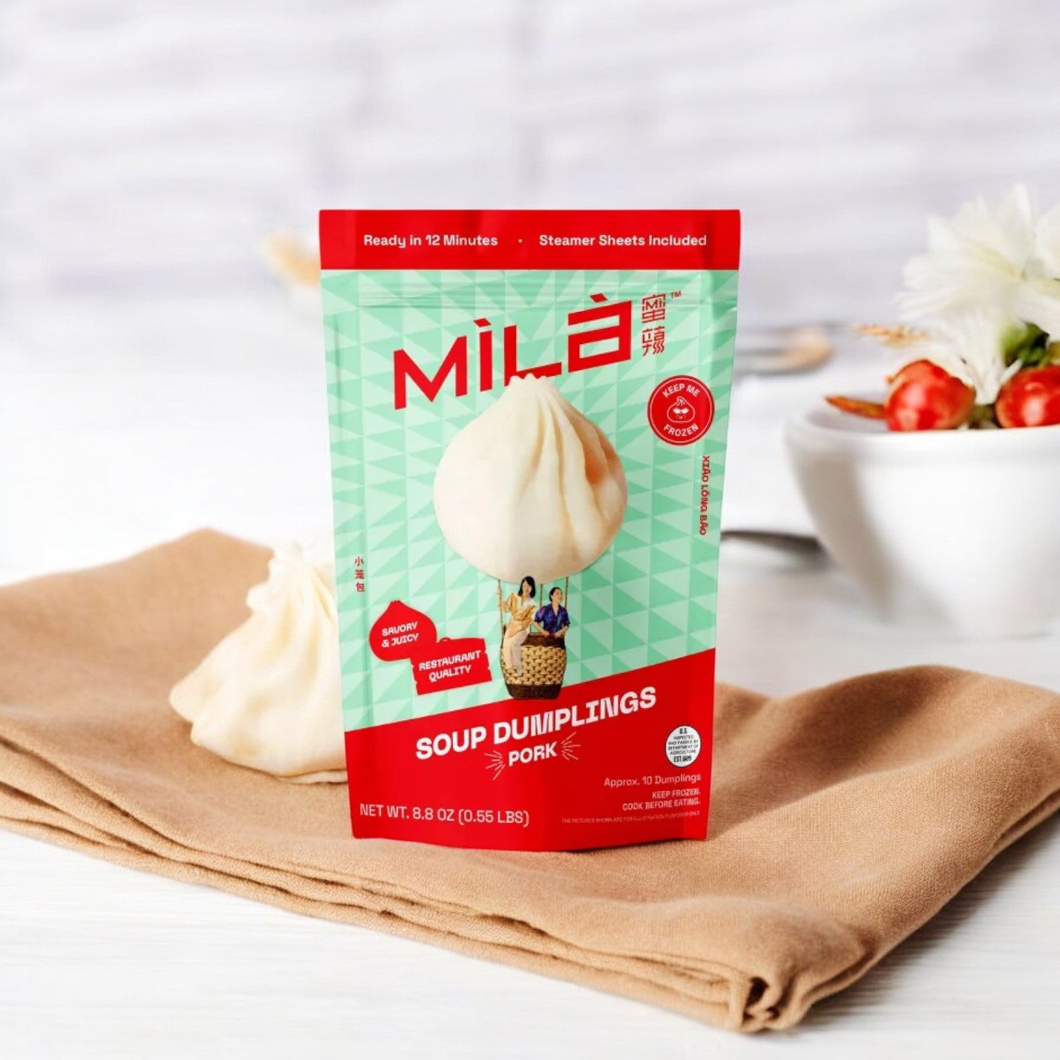 A pack of MiLa Pork Soup Dumplings, Frozen 8.8 oz, is elegantly displayed on a napkin with one dumpling artfully positioned beside it. The arrangement is complemented by flowers in the background, showcasing the fusion of authentic Chinese cuisine with the ease of modern frozen convenience.