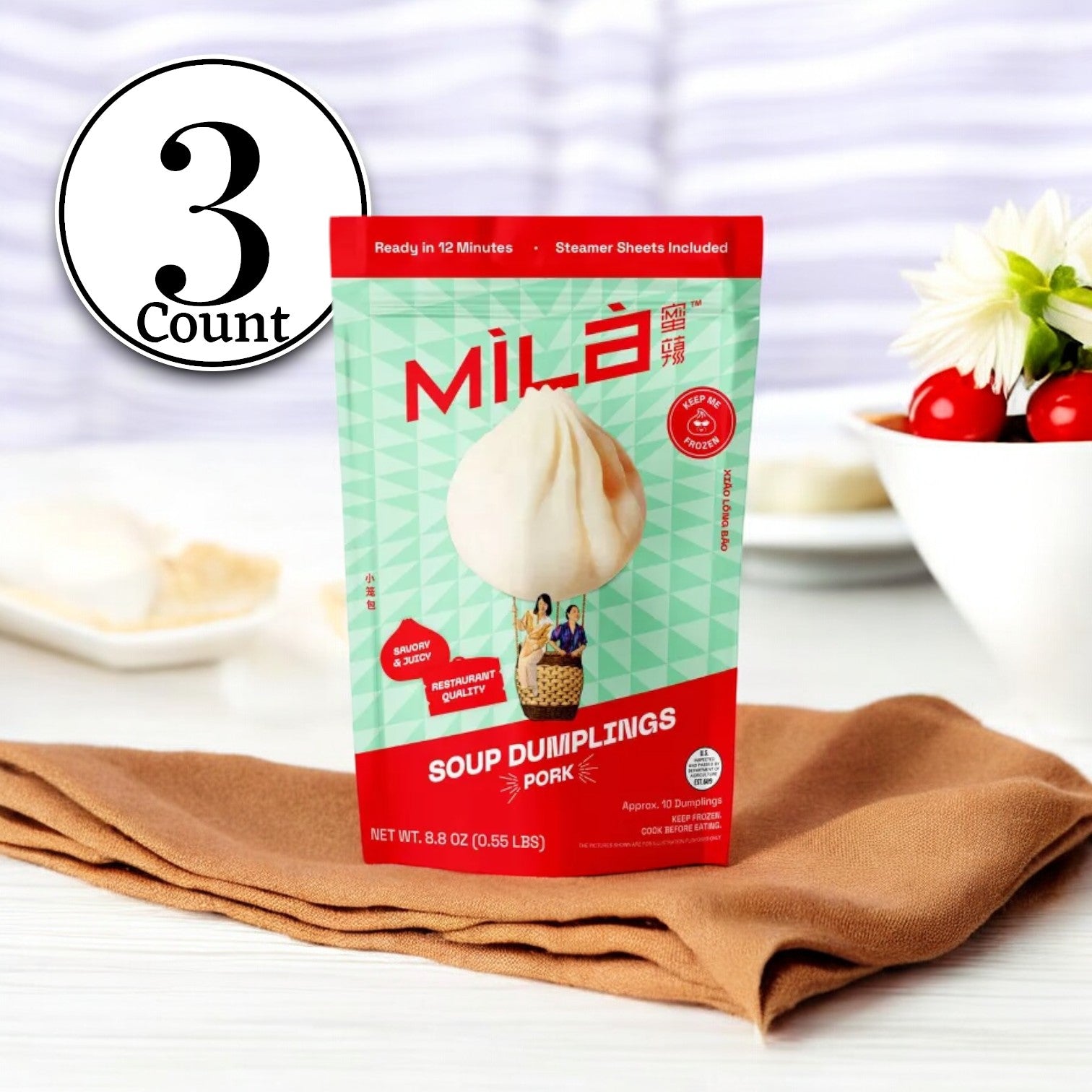 A package of MiLa Pork Soup Dumplings, representing the essence of authentic Chinese cuisine, is placed on a table with a brown napkin and a bowl of vegetables in the background. The packaging proudly displays "10 Count" and offers frozen convenience, boasting that they are "Ready in 12 Minutes.