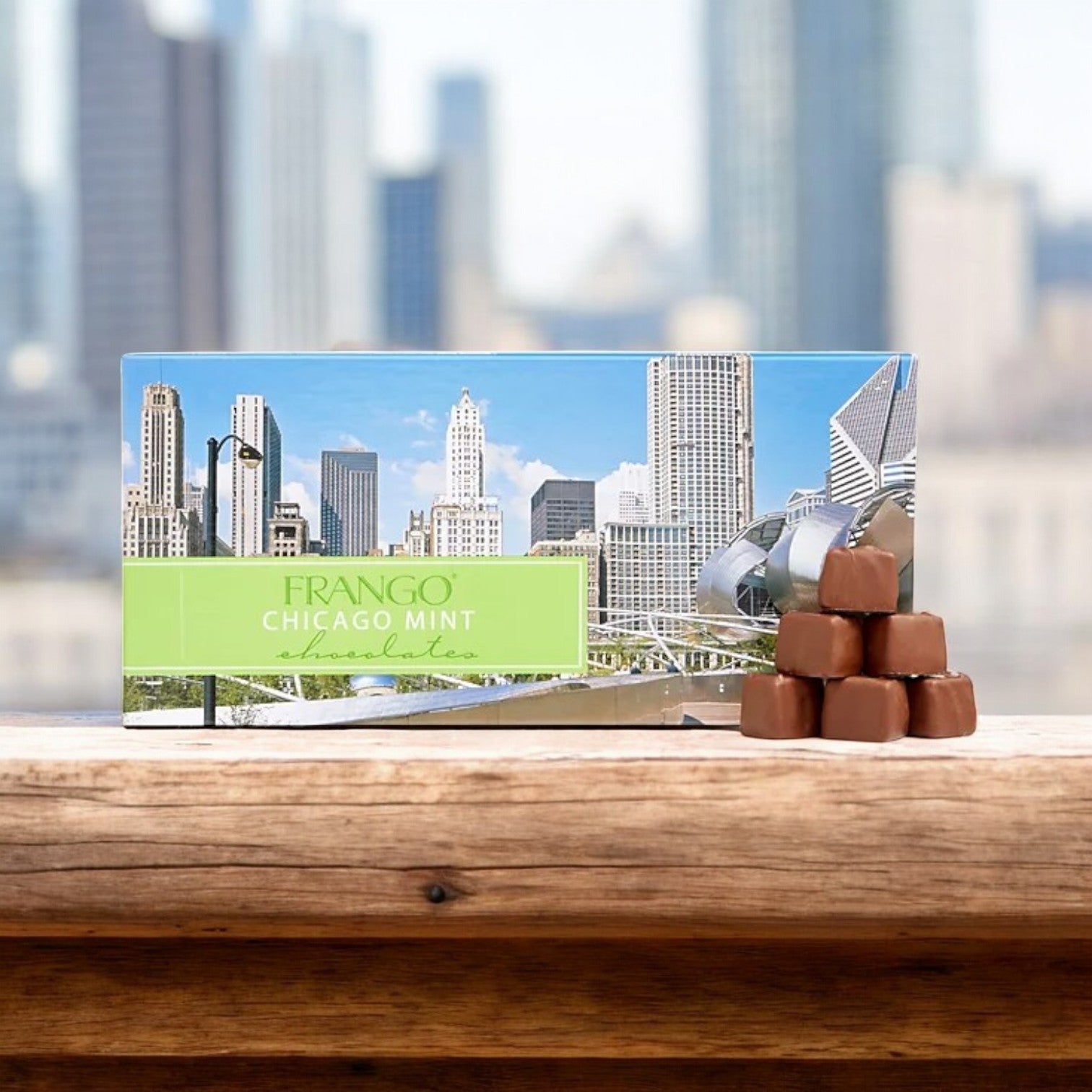 A box of Frango mint chocolates and Gino's East Pizza combo rests on a wooden surface, with four chocolates stacked in front against a backdrop of a city skyline and modern architecture, capturing the essence of Chicago like its famed deep-dish pizza.