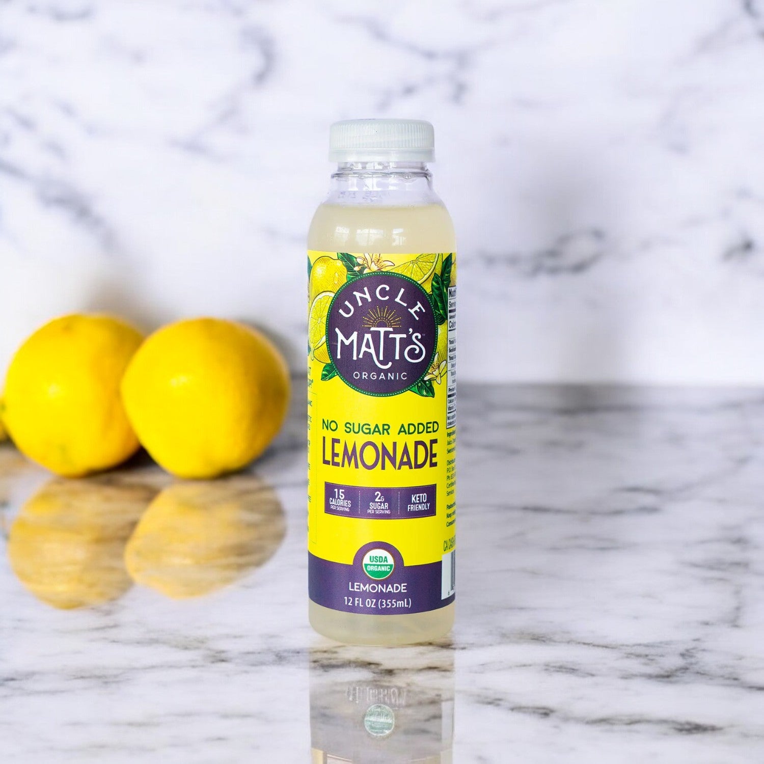 A 12 oz bottle of Uncle Matt's Organic Lemonade is placed on a marble countertop, crafted with organic lemons and organic stevia, with two fresh lemons in the background.