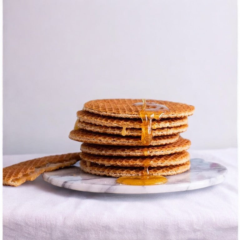 A stack of four Daelmans Individually Wrapped Caramel Stroopwafels with caramel dripping down the sides is placed on a marble surface. One broken stroopwafel lies beside the stack.