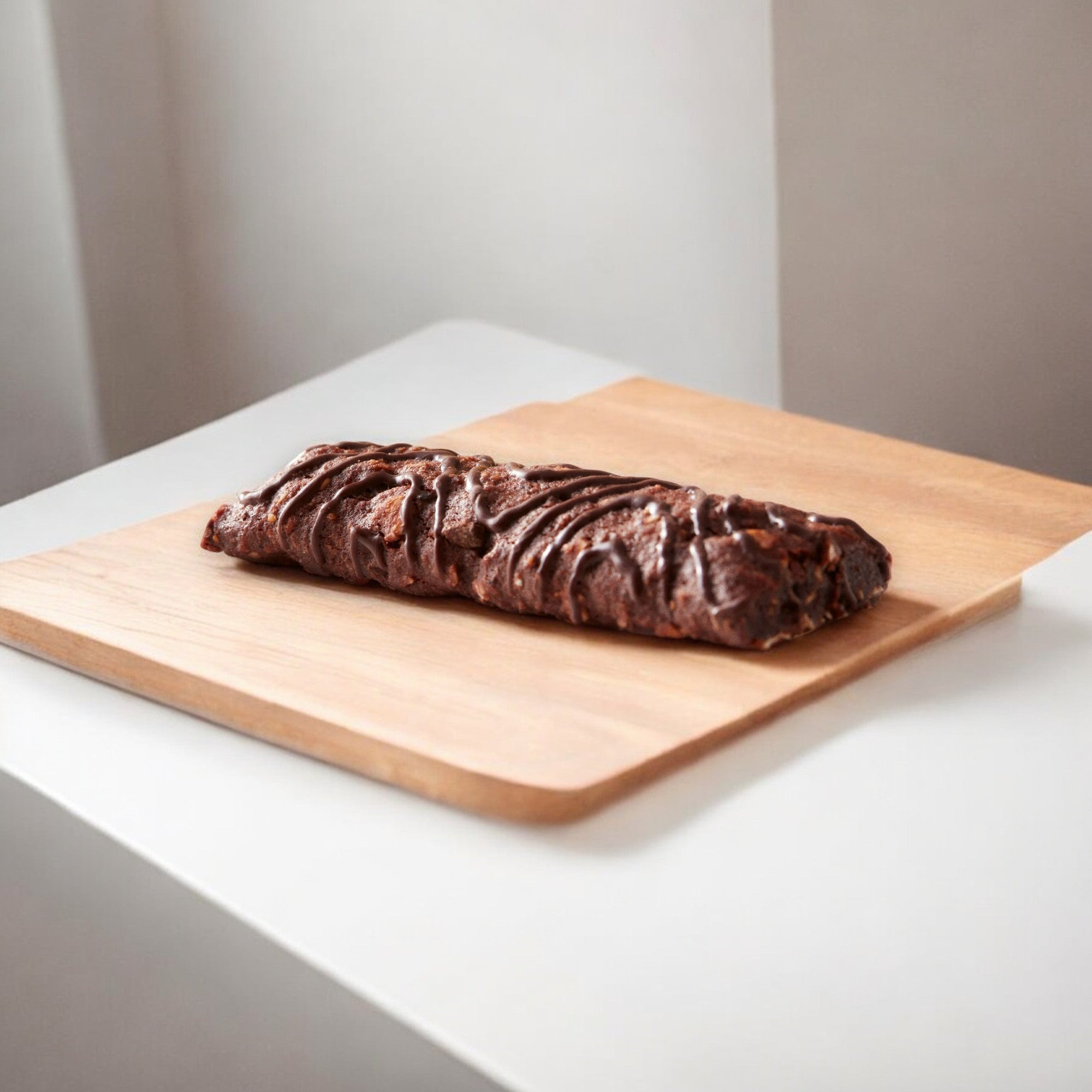 A Clif Zbar Chocolate Brownie Soft Baked Whole Grain Snack Bar, weighing 1.27 oz, rests on a light wooden cutting board placed on a white surface, offering an artisanal touch reminiscent of whole grain nutrition found in the Clif Bar brand.