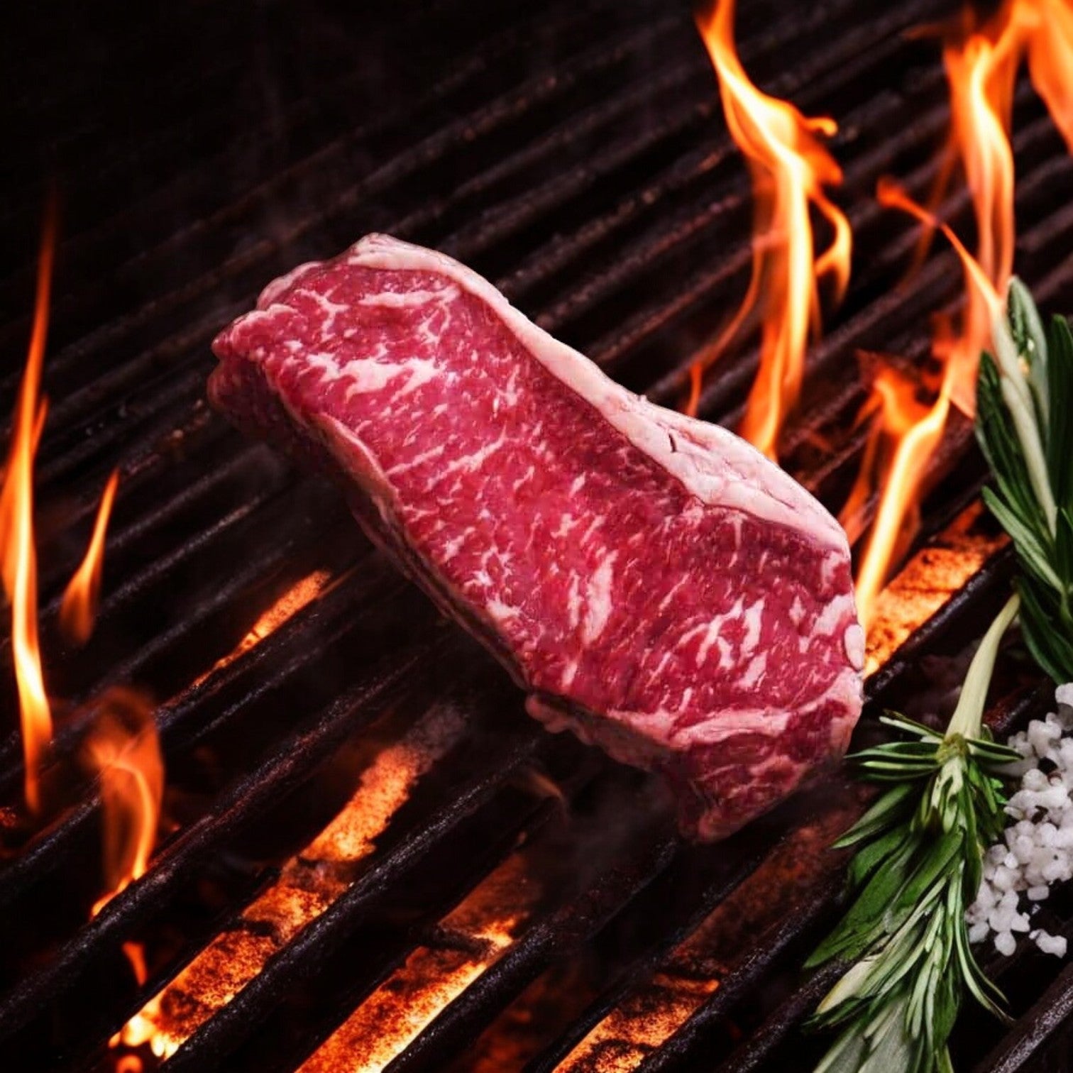 A Mino Prime Steaks – One, 16 oz Wet-Aged New York Strip sizzles on the grill, surrounded by dancing flames, rosemary sprigs, and salt crystals.