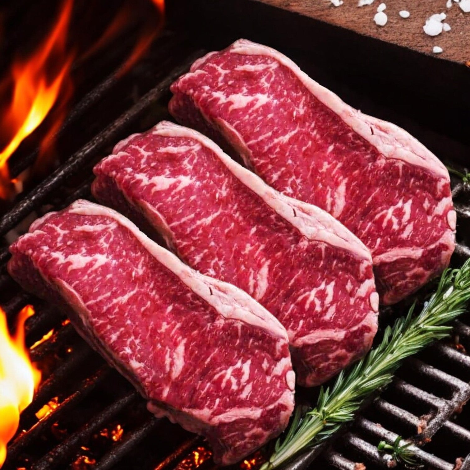 Three cuts from the Mino Prime Steaks - Top Seller Box, featuring a premium Wet-Aged New York Strip, sizzle on the grill as flames dance around them, with rosemary and coarse salt enhancing the aroma.