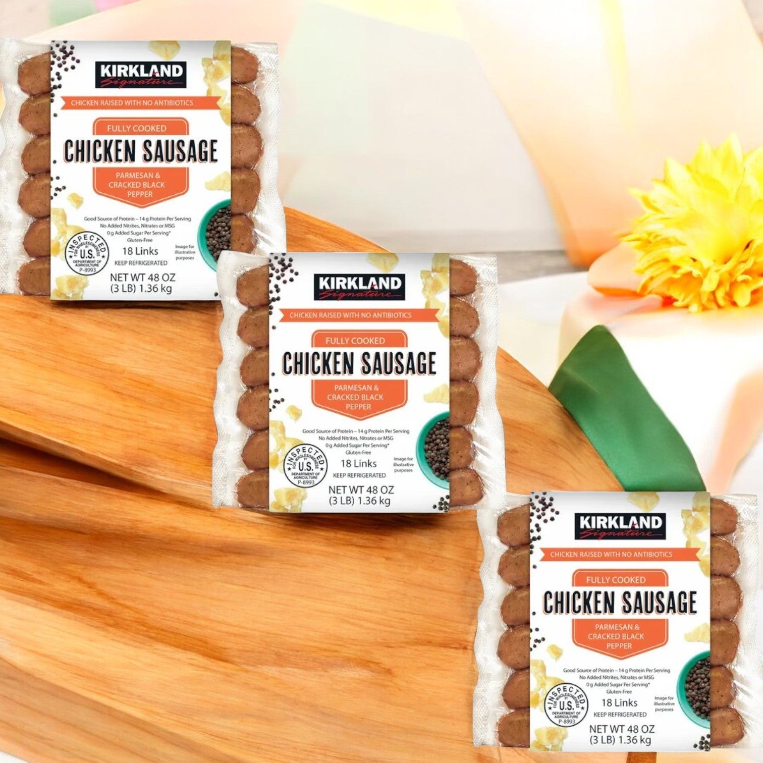 Three packages of Kirkland Signature Parmesan Black Pepper & Parmesan Chicken Sausage are placed on a wooden surface, highlighted by a decorative flower in the background.