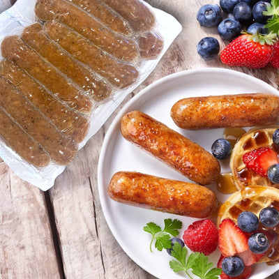 A plate featuring Amylu Chicken Sausage Links, Apple & Maple with a waffle topped with syrup and berries, garnished with parsley; an uncooked sausage pack showcasing the all-natural ingredients rests beside it.