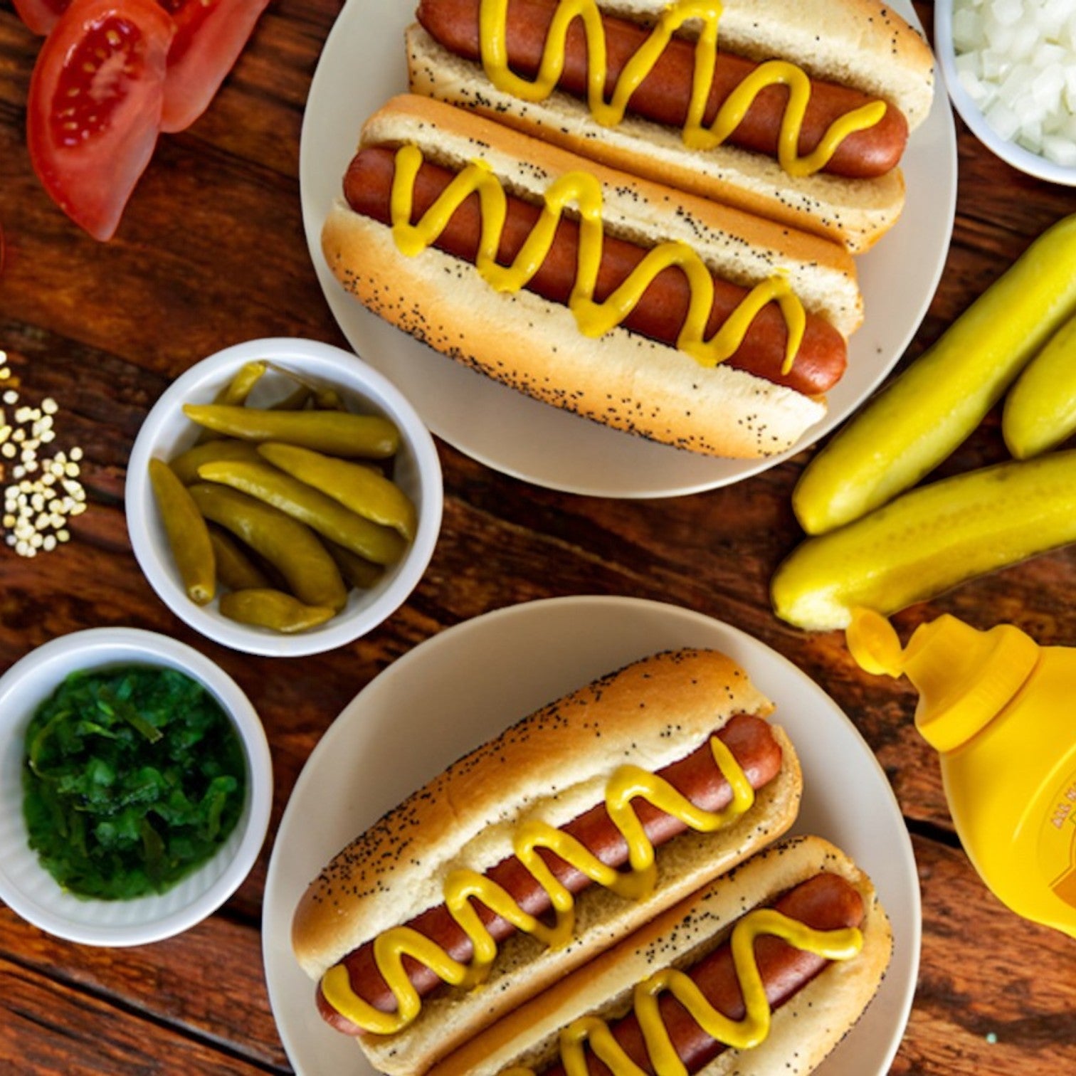 On a wooden table, Vienna Beef 6" Skinless Polish Sausages are topped with mustard on two plates, surrounded by bowls containing toppings like pickles, chopped onions, and tomatoes.