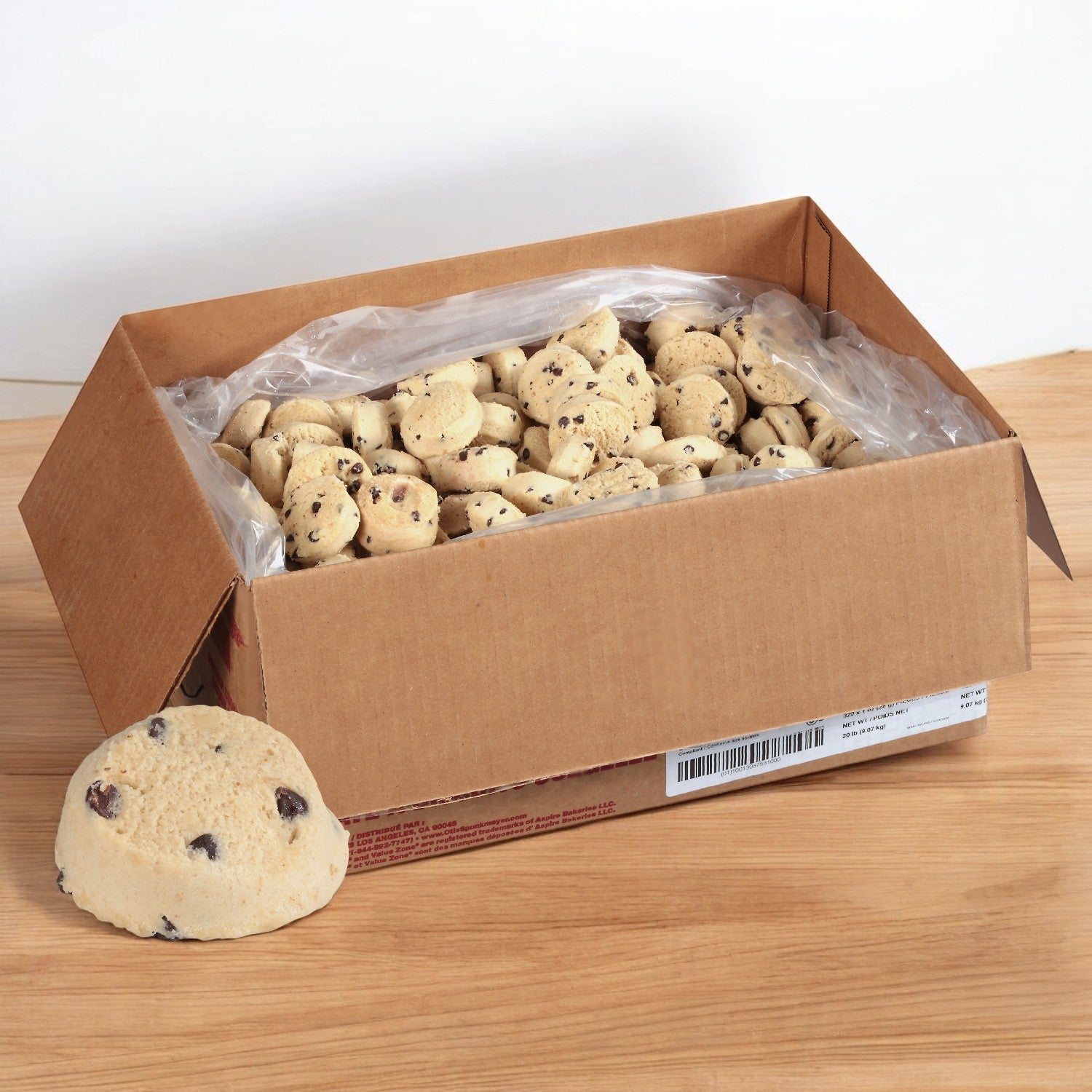 A cardboard box of Otis Spunkmeyer Dough Cookie Chocolate Chip, 320/1 oz., sits open on a wooden surface, with bulk premium dough pieces next to one individual piece, capturing the essence of irresistible Otis Spunkmeyer Chocolate Chip Cookies.