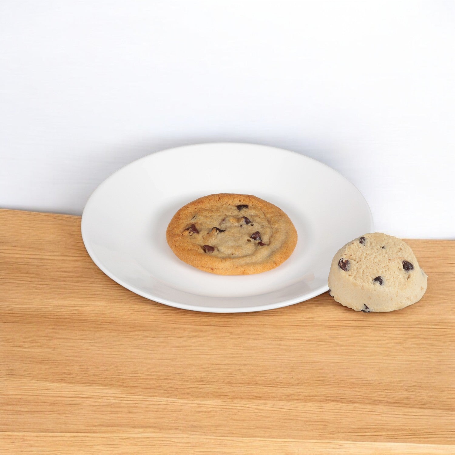 An Otis Spunkmeyer Dough Cookie Chocolate Chip, baked to perfection, sits on a white plate next to an unbaked premium dough ball on a wooden surface.