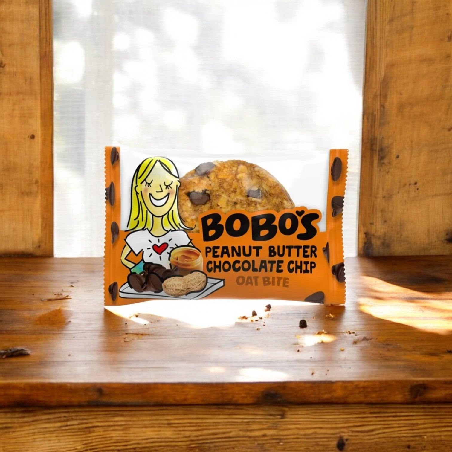 A package of Bobo's PB & Chocolate Chip Oat Bites, 1.3 oz -1 Count, showcasing wholesome ingredients, rests on a wooden surface with crumbs scattered around it, set against a sunlit window backdrop.