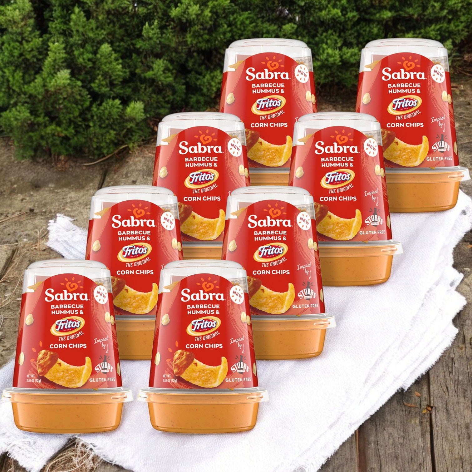 Eight packs of Sabra BBQ Hummus Dip with Fritos, 2.55 oz each, are stacked on a white cloth outdoors, creating an ideal portable snack amidst greenery.