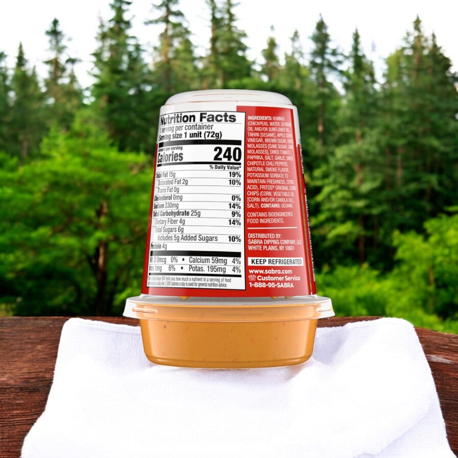 A Sabra BBQ Hummus Dip with Fritos from the 8 pack (2.55 oz each) rests on a white napkin, with pretzels, creating an ideal portable snack against green trees; the nutrition label is visible.