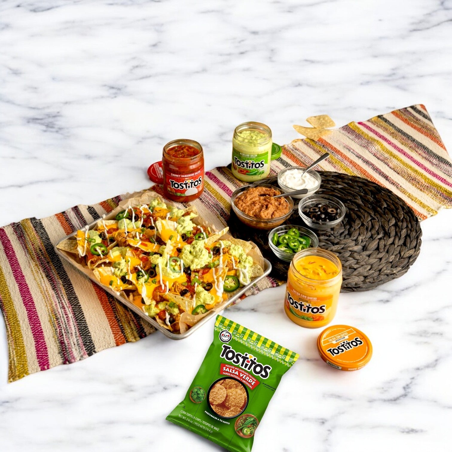 A platter of Tostitos Salsa Verde Tortilla Rounds, topped with cheese and jalapeños, is ready for snack enthusiasts. Around it are Tostitos salsa, guacamole, and queso dips on a woven mat over marble—ideal for dipping and snacking joy.