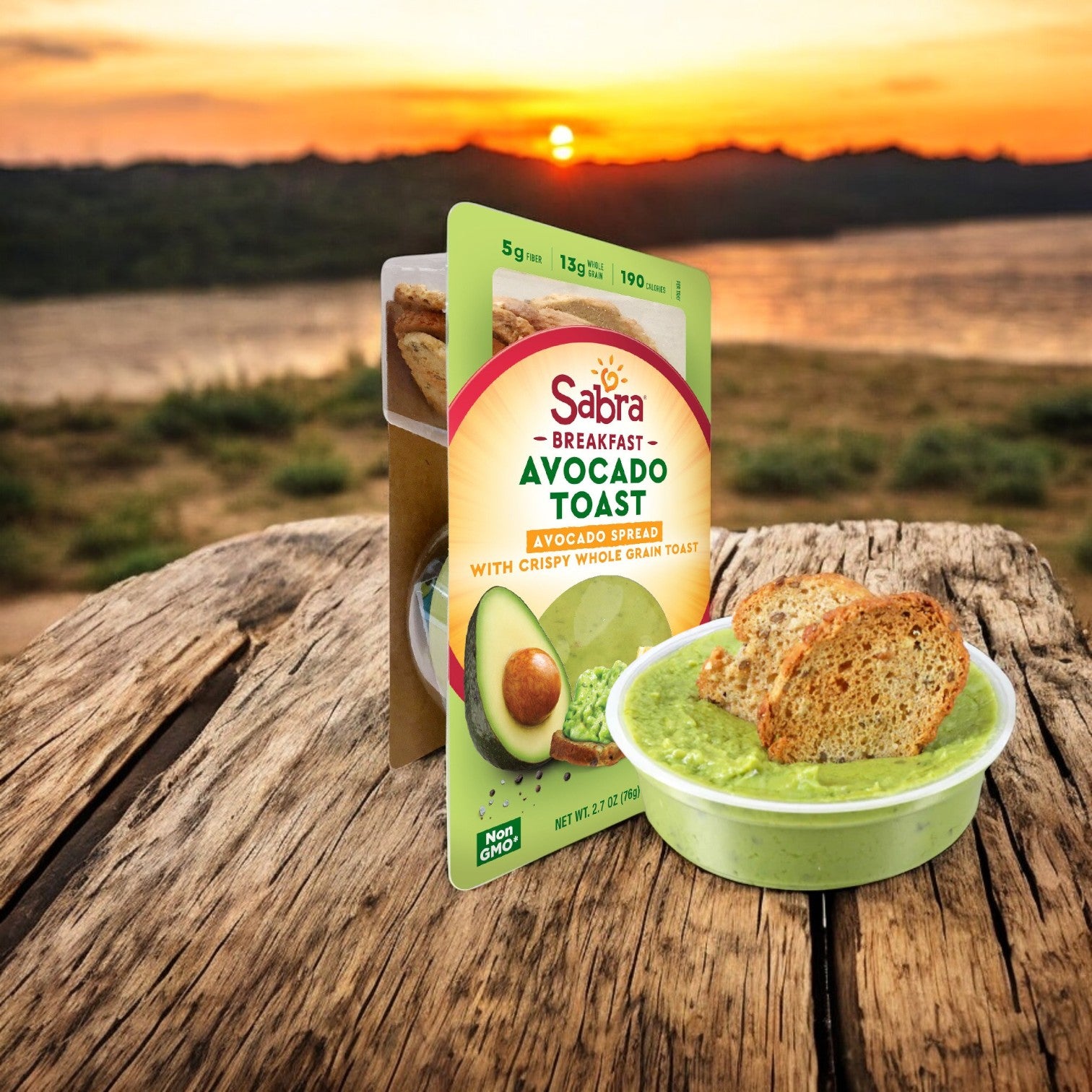 Sabra Avocado Chunky Spread with Toast, an 8-count pack at 2.7 oz each, is placed on a wooden surface near a lake sunset, offering an authentic flavor experience.