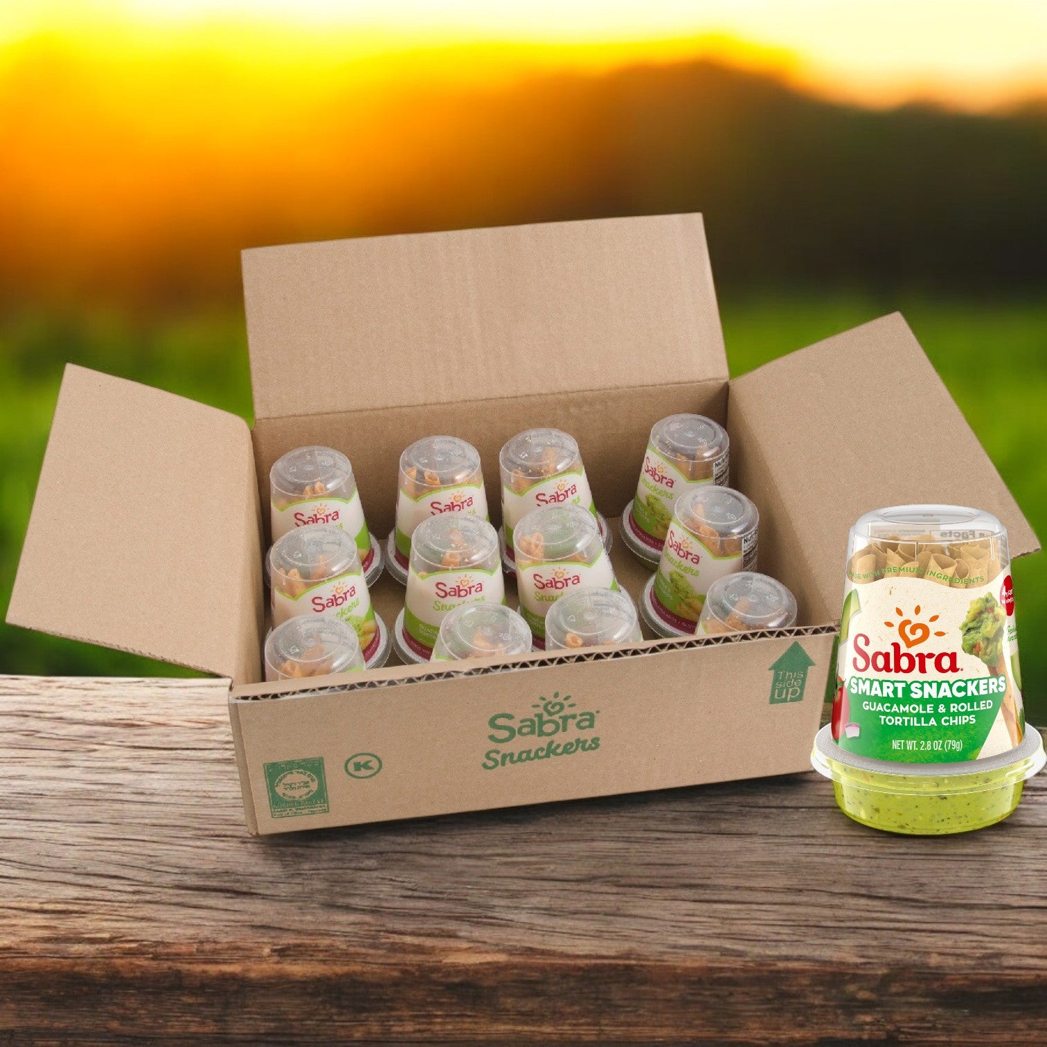 A box of Sabra Guacamole Fresh with Tostitos Grab n' Go (12 pack, 2.8 oz each) offers a delightful portable snack. One package sits on a wooden surface against a sunlit greenery backdrop.