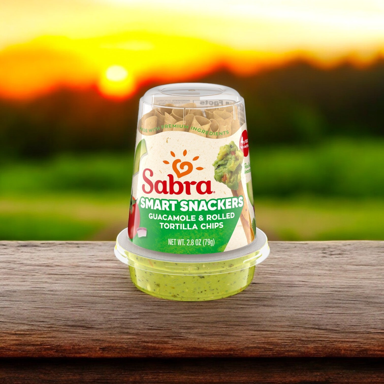 The Sabra Guacamole Fresh with Tostitos Grab n' Go, 2.8 oz package sits on a wooden surface, inviting you to savor this portable snack amid a scenic sunset and lush green hills.