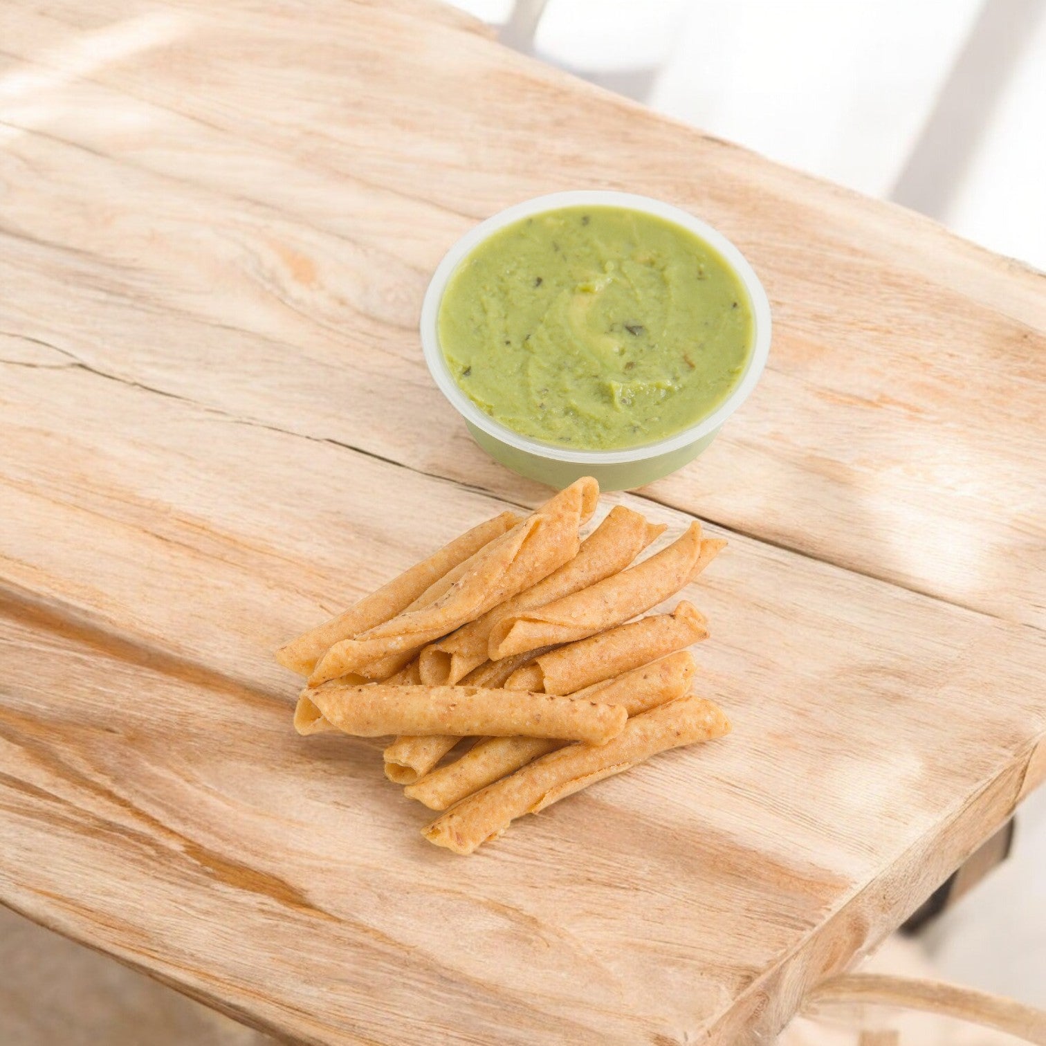 A small bowl of Sabra Guacamole Fresh with Tostitos sits next to crispy rolled tacos on a wooden surface, offering a delightful contrast to vibrant Classic Hummus, perfect as a portable snack for any occasion.