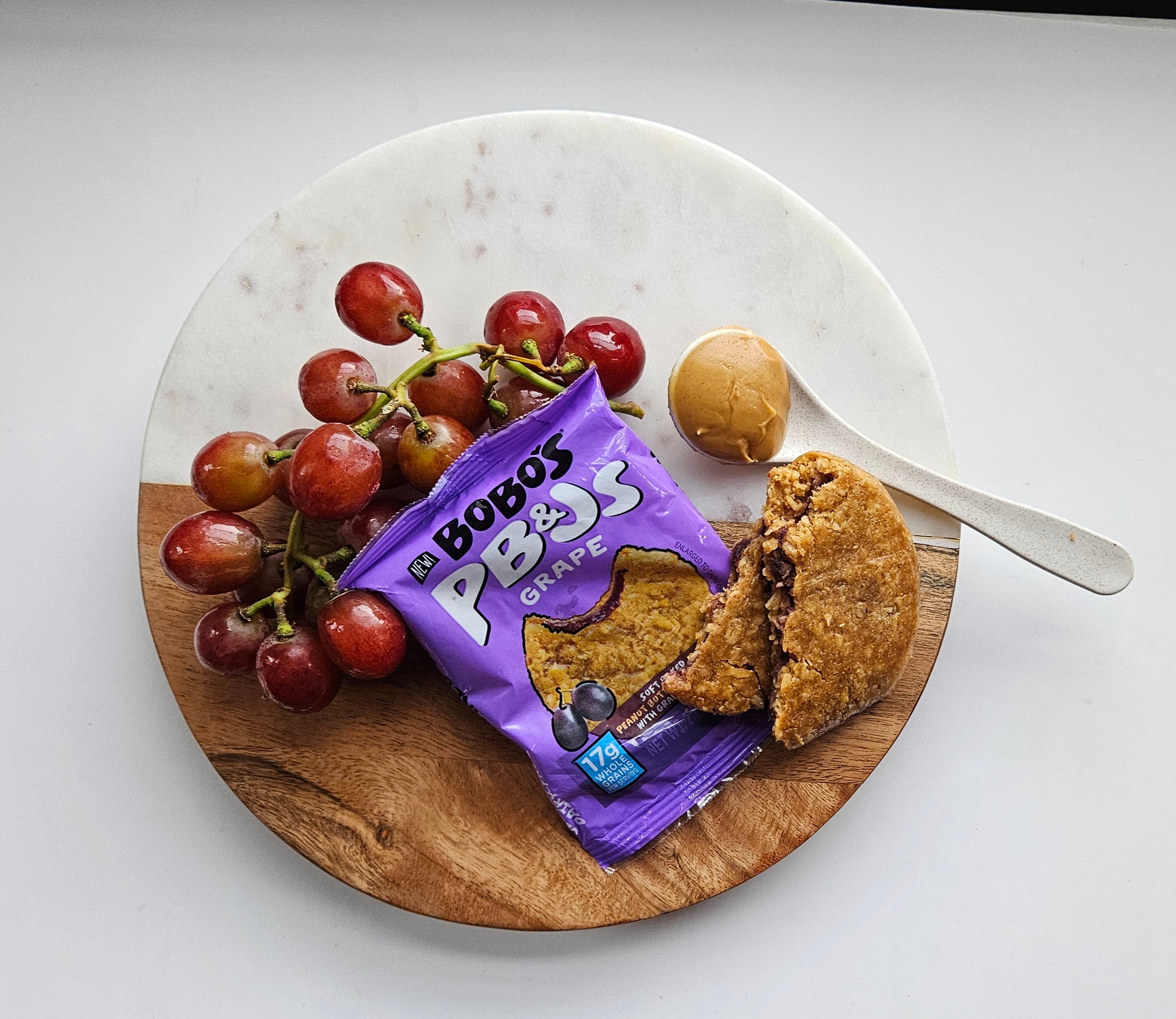 A snack plate featuring red grapes, a packet of Bobo's PB&Js Grape, 2.1 oz - 1 Count—a perfect vegan treat—an opened Bobo's pastry, and a spoonful of peanut butter on a white and wooden round board.