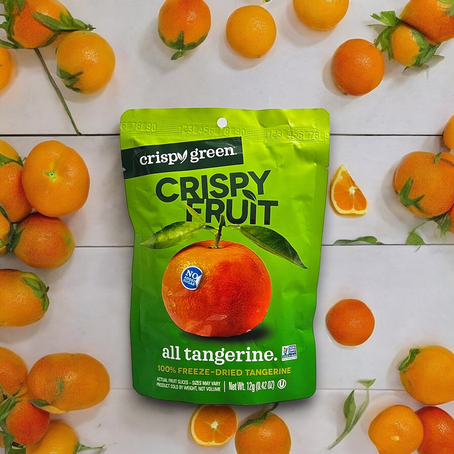 A green bag of Crispy Green Crispy Fruit All Tangerine-0.35 oz-1 Count is surrounded by fresh tangerines. The package reads "Crispy Fruit," "all tangerine," "100% freeze-dried tangerine," and "Net Wt. 0.35oz (10g). Try also our Crispy Green Pineapple Slices for a real pineapple slice experience, perfect for any occasion!