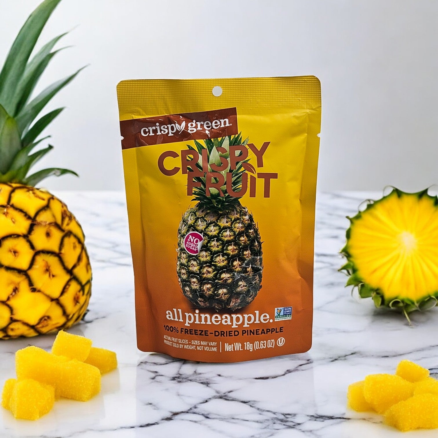 Packaging of Crispy Green Crispy Fruit, All Pineapple (0.35 oz, 1 Count) on a marble surface, surrounded by a whole pineapple, real pineapple slices, and yellow pineapple cubes, showcasing an environmentally conscious choice.