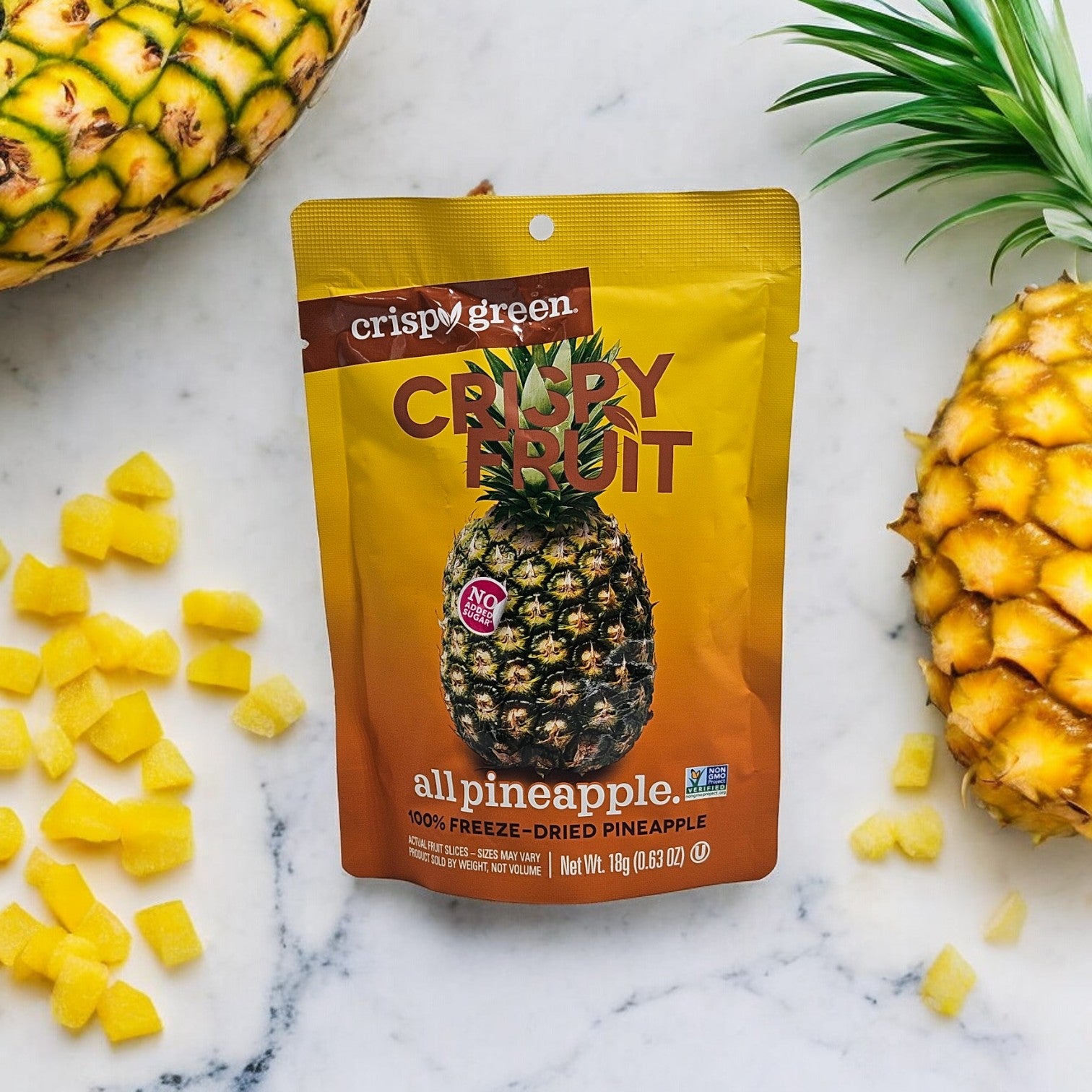 A bag of Crispy Green Crispy Fruit, All Pineapple-0.35 oz-1 Count is placed on a marble surface, surrounded by cut pineapples and pineapple cubes. The yellow and dark brown packaging features an image of real pineapple slices, reflecting Crispy Green's environmentally conscious approach to preserving natural flavors.