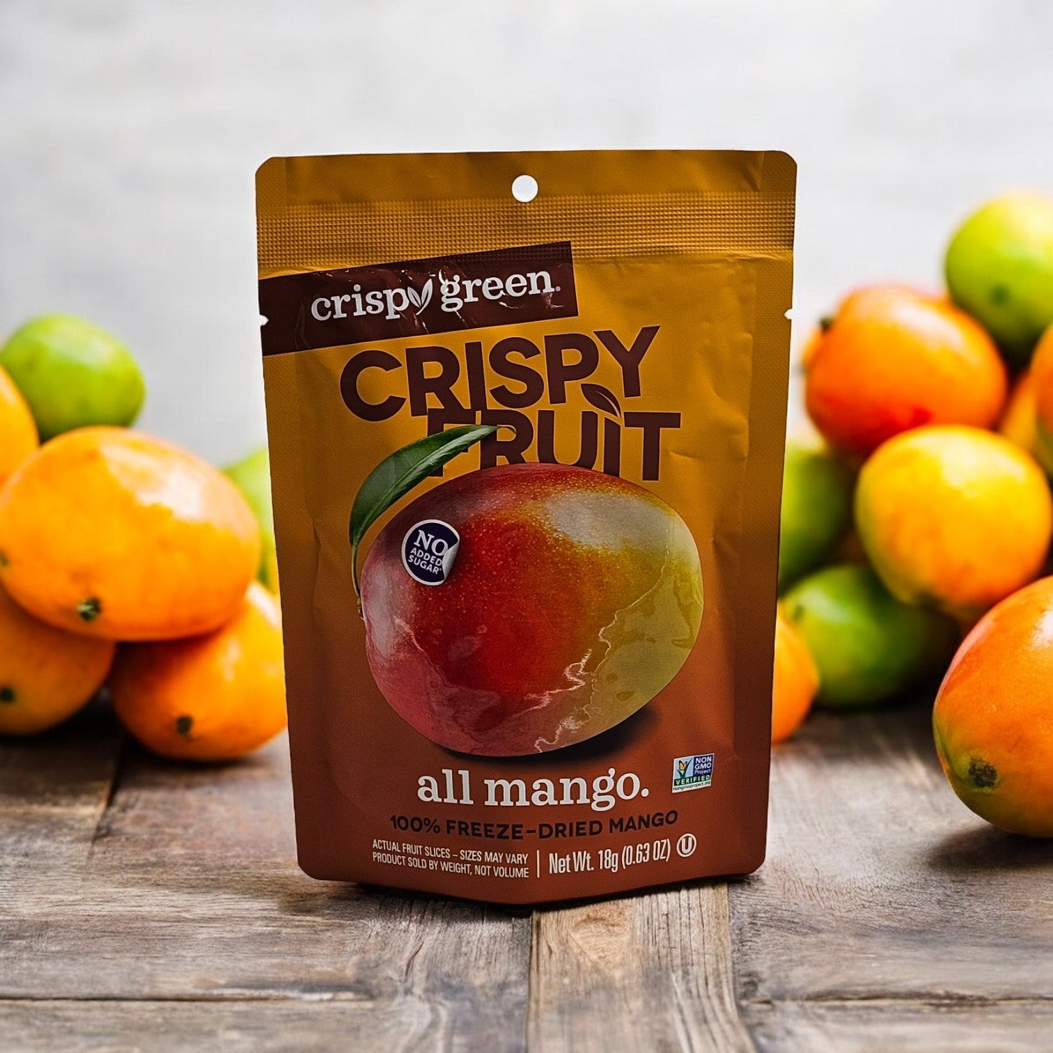 A bag of Crispy Green Crispy Fruit All Mango - 0.35 oz - 1 Count snacks is displayed on a wooden surface with fresh mangoes in the background. The bag features a large mango image and various product labels, highlighting Crispy Green's commitment to sustainable snacks.