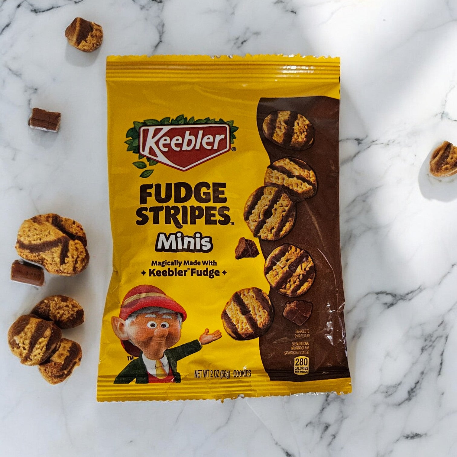 A yellow pack of Keebler Fudge Strips-2 oz - 1 Count is displayed on a marble surface, surrounded by a few cookies and chocolate pieces, showcasing the genuine Keebler fudge and buttery shortbread that make these treats irresistible.