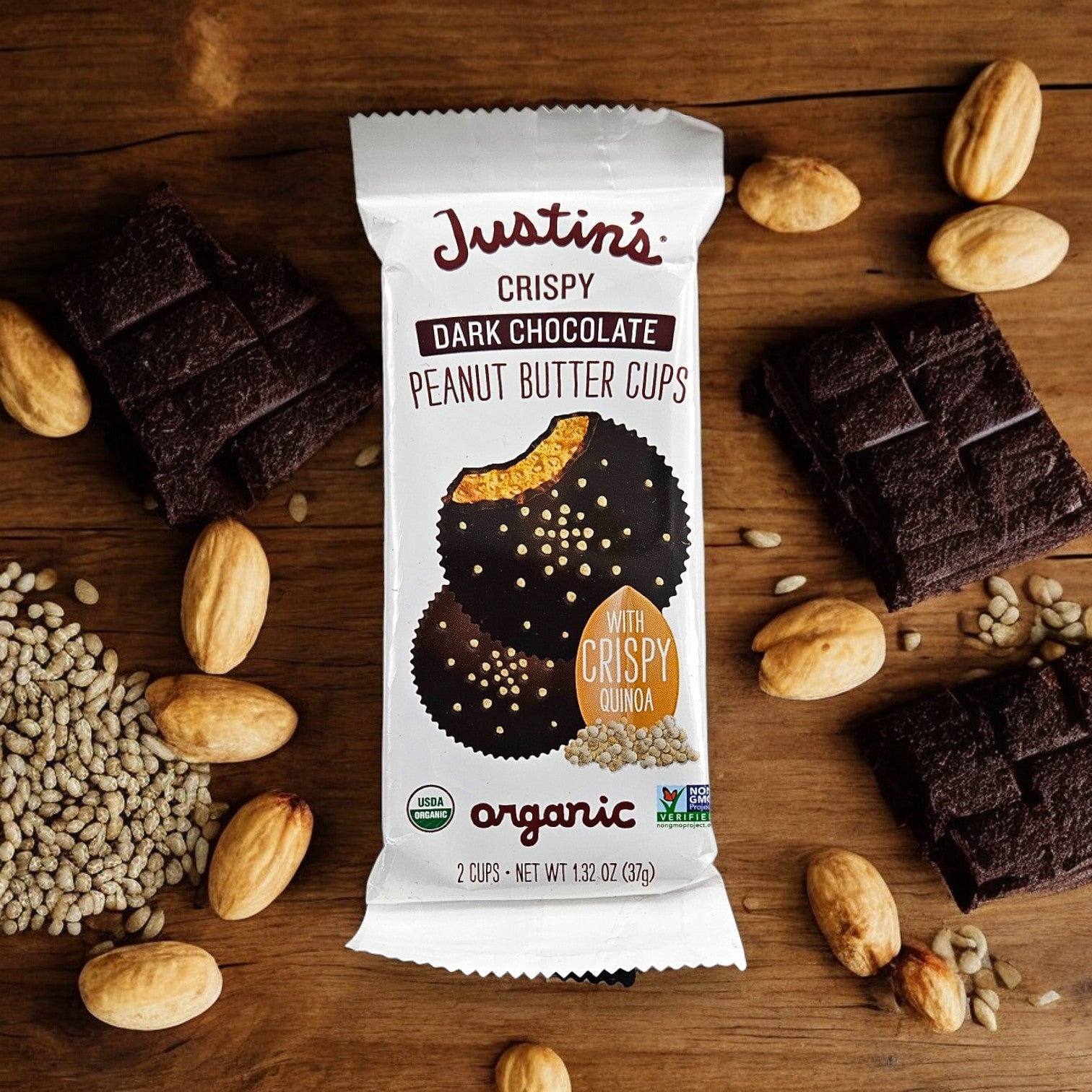 A 1.4 oz package of Justin's Organic Dark Chocolate Peanut Butter Cups, surrounded by peanuts, whole and crushed chocolate, and grains. The packaging highlights that the product is organic and includes crispy quinoa.