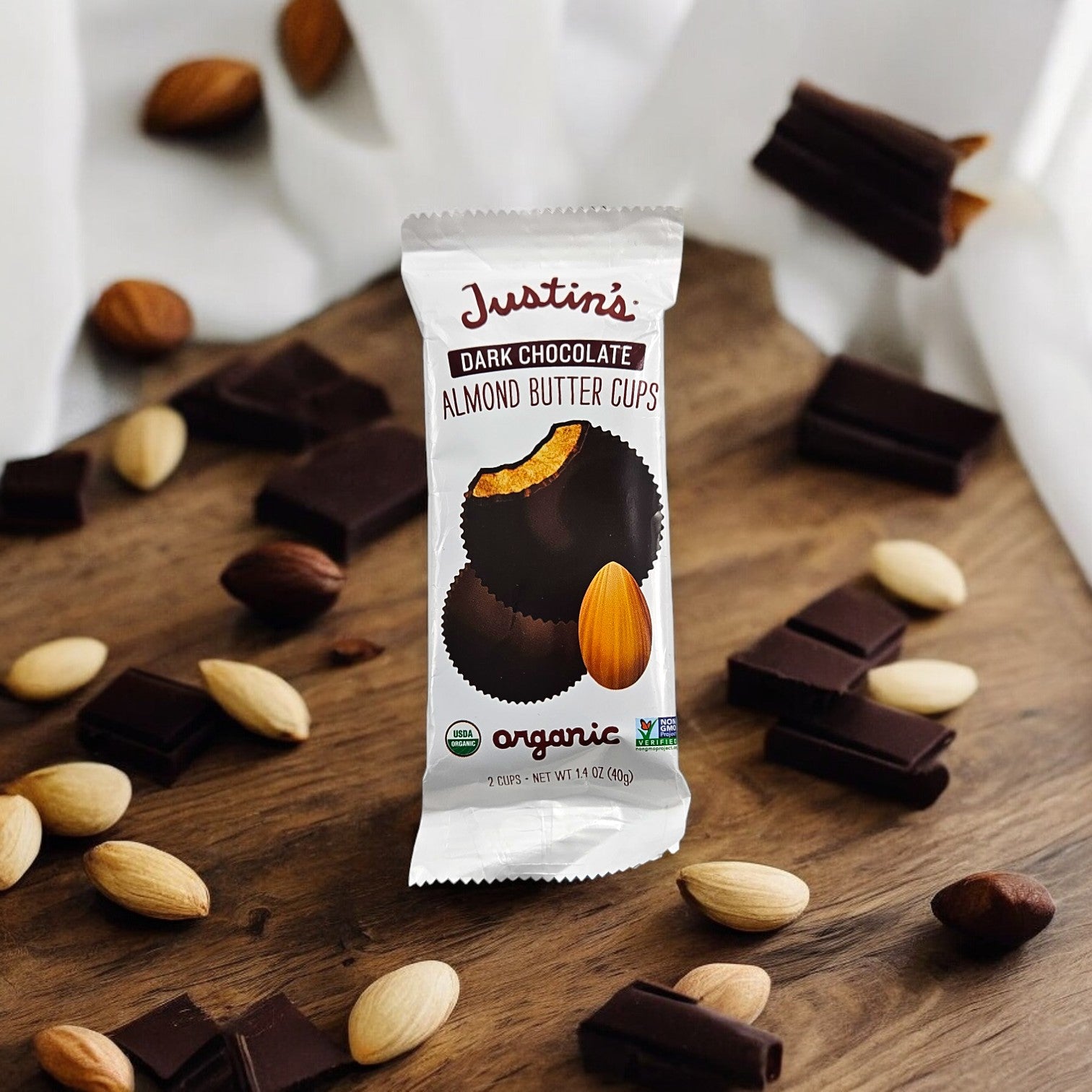 A package of Justin's Organic Dark Chocolate Almond Butter Cups (1.4 oz, 1 Count) is placed on a wooden surface surrounded by almonds and pieces of dark chocolate.