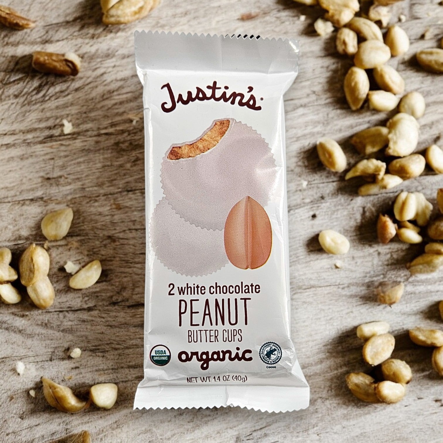 A package of Justin's Organic 2 White Chocolate Peanut Butter Cups (1.4 oz, 1 Count), made with the finest organic ingredients, surrounded by scattered peanuts on a wooden surface.