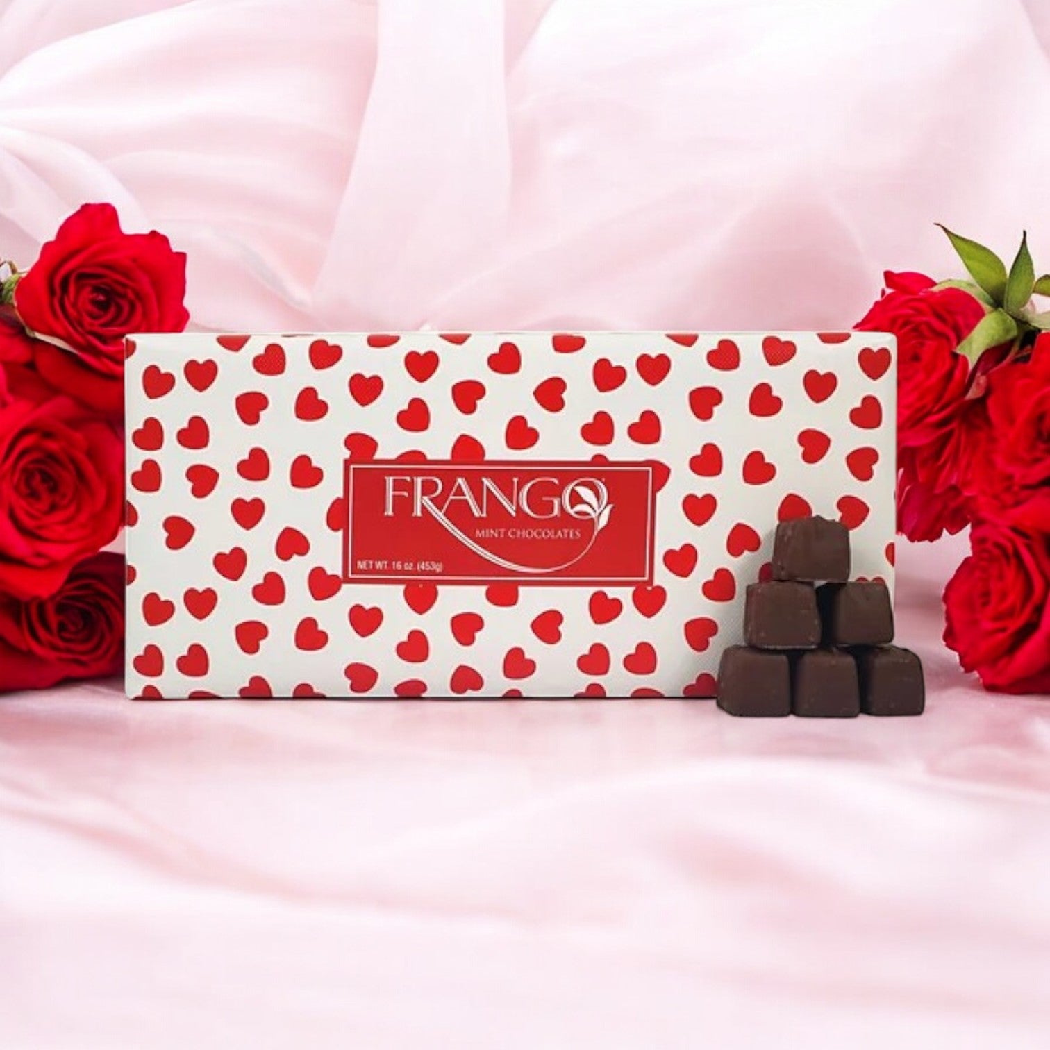 A box of Macy's Frango Chocolates 1 LB Valentine's Heart Wrapped Milk Mint rests on pink fabric, alongside red roses and stacked milk chocolate pieces. Ideal for Valentine's Day.