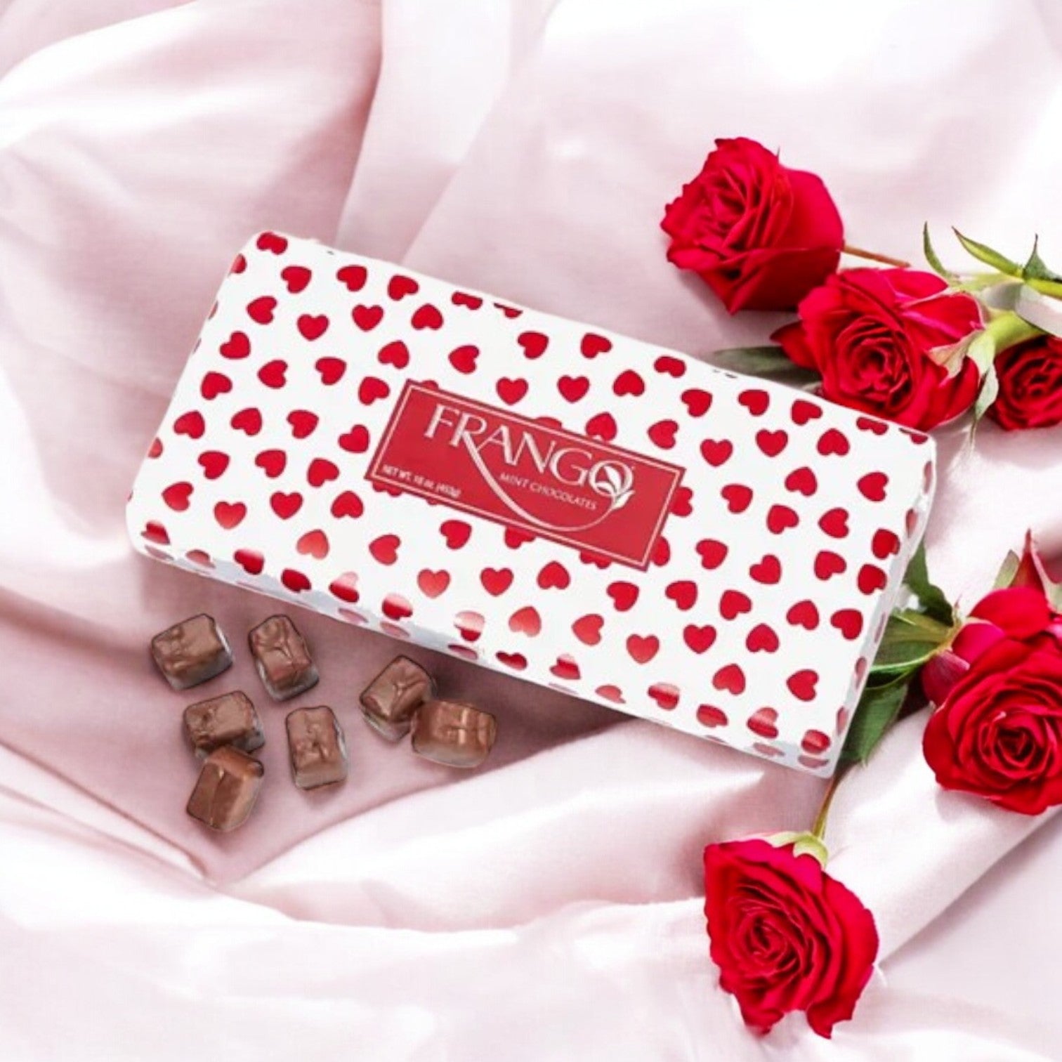 A Frango Chocolates 1 LB Valentine's Heart Wrapped Milk Mint Box by Macy's sits on pink fabric, surrounded by red roses and unwrapped milk chocolates, ideal for a Valentine's Day treat.