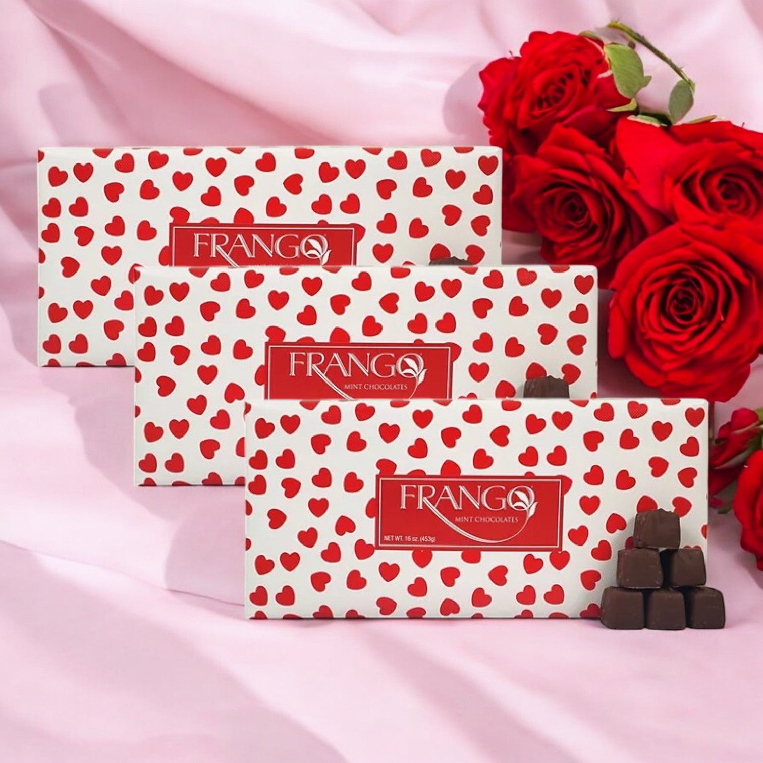 Three boxes of Macy's Frango Chocolates 3 LB Valentine's Heart Wrapped Milk Mint Box of Chocolates are displayed on a pink surface beside several red roses and stacked chocolate pieces.