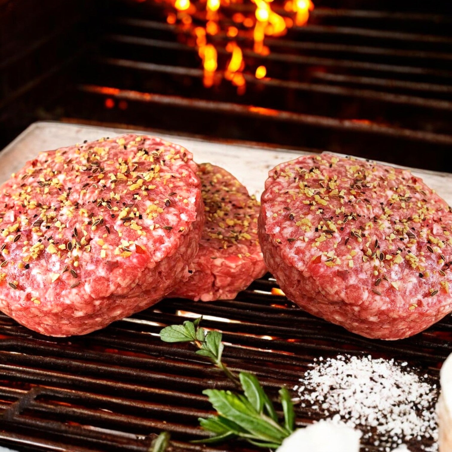 Two Mino Prime Steaks USDA Prime Steak Burgers grill with subtle flames, surrounded by salt and fresh herbs, highlighting their premium quality.